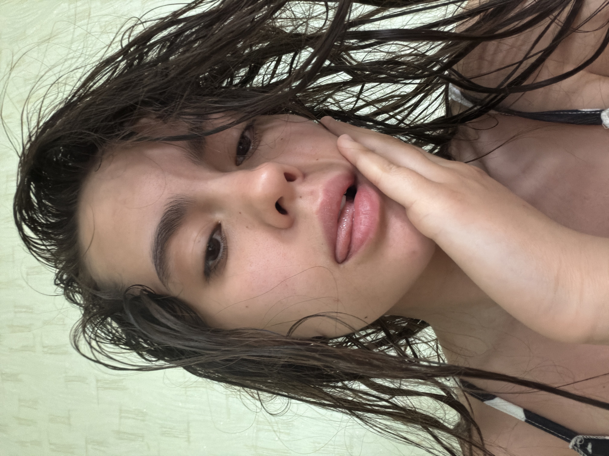 A close-up, rotated, high-angle shot depicts a female with wet, dark brown hair. Her skin is fair, and she is touching her cheek with her right hand. Her tongue is slightly extended between her lips. The background shows a light green, patterned surface. The setting is not explicitly identifiable as Chaikovsky, Russia, and there is no other evidence about the location.