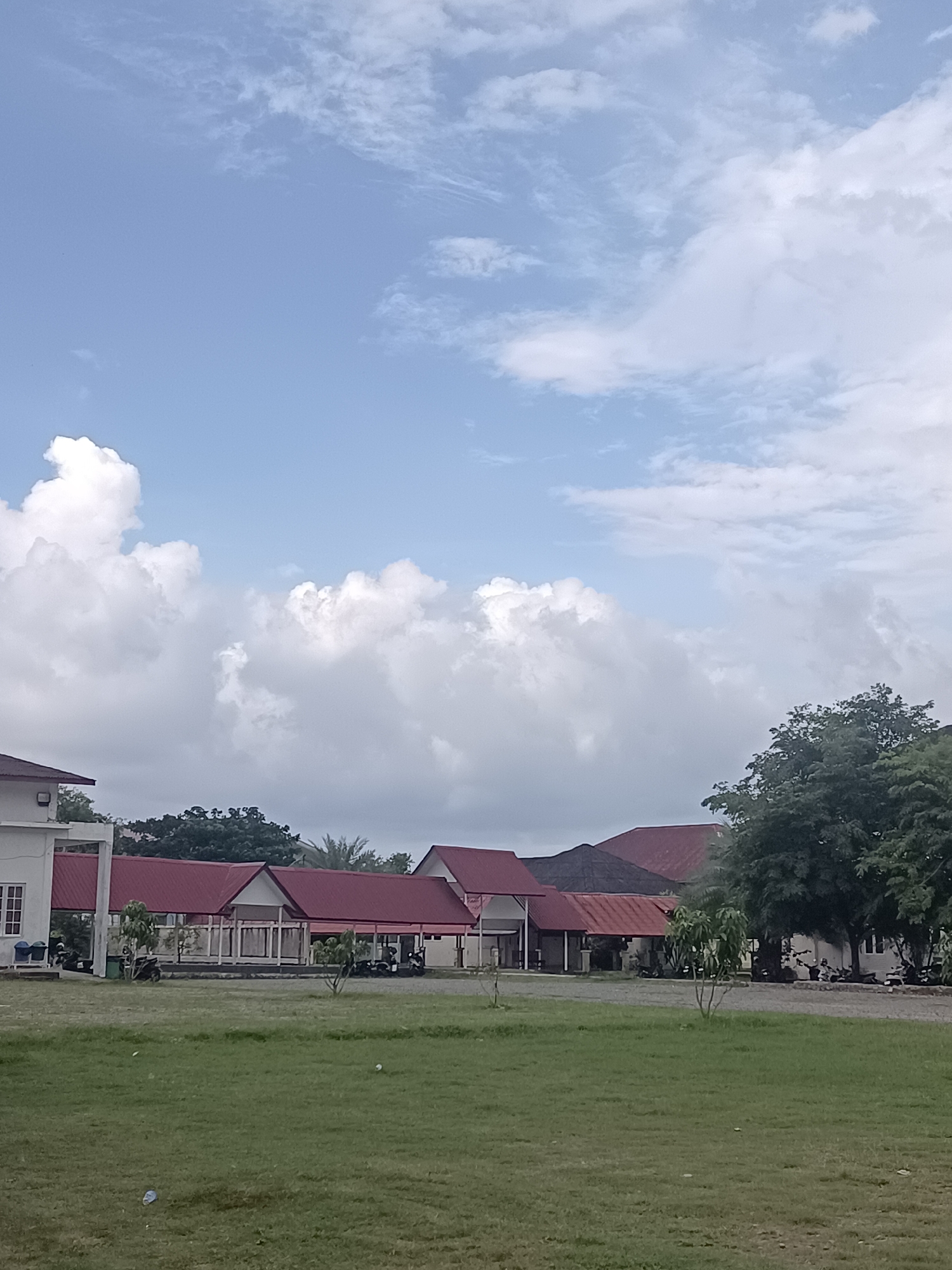 This image depicts a serene outdoor scene in Mesjid Reuntoh, Indonesia. The foreground is dominated by a lush, green grassy field, with a few scattered white pieces of litter. In the middle ground, a row of buildings with distinctive red roofs stretches across the frame. These structures appear to be single-story with open-air covered walkways. To the left, a white building with visible windows and an exterior light fixture stands out. The buildings are flanked by trees and vegetation, with a large, mature tree on the right side of the image providing a strong vertical element. Several motorcycles are parked in front of the buildings, suggesting a place of activity or residence. The sky above is a prominent feature, a bright blue filled with a dramatic display of white, fluffy cumulus clouds. Some of these clouds are large and voluminous, casting subtle shadows, while others are wispy and spread out. The overall impression is one of a calm, perhaps early morning or late afternoon, with pleasant weather. There is no visible text in the image.