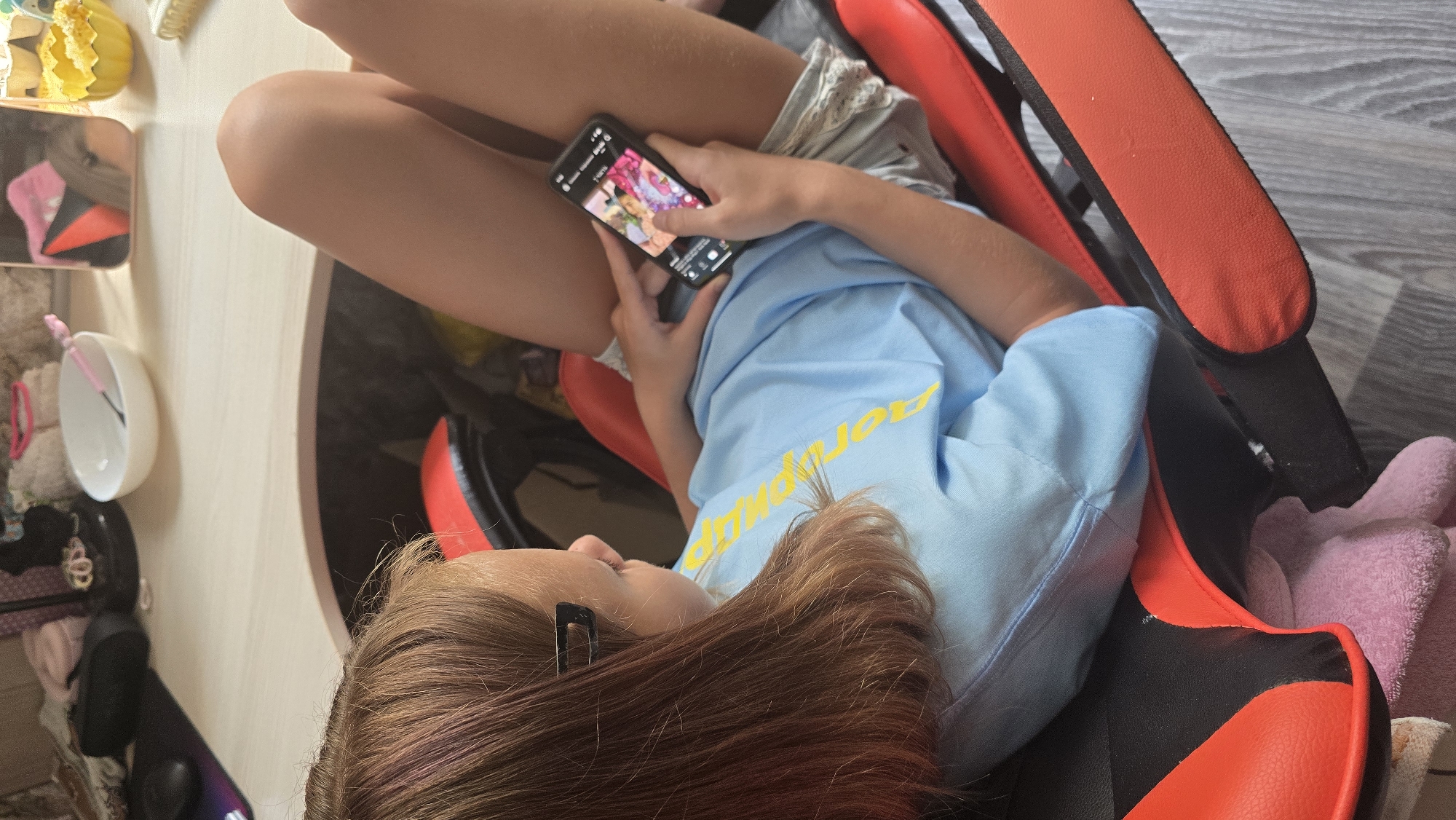 A young girl with shoulder-length brown hair, with subtle pink highlights, is seated in a black and red gaming chair. She is wearing a light blue t-shirt with yellow lettering and grey shorts with white trim. She is holding a smartphone in her hands, looking at its screen. The phone displays a colorful image. In the background, a wooden desk is visible with various items including a white bowl with a pink utensil, a black computer mouse, and other indistinct objects. The setting appears to be an indoor environment, likely a room within a residence in Novohuivynske, Ukraine.