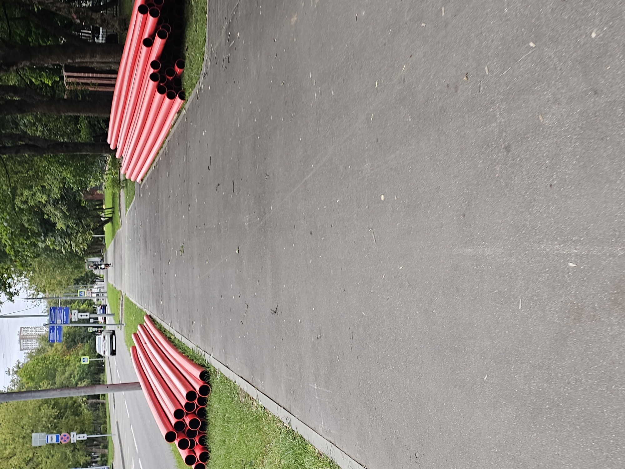 Two stacks of bright red, large-diameter pipes are laid on the grass next to a paved pedestrian walkway. The pipes are roughly cylindrical and appear to be made of plastic, likely for underground utility installation. The walkway itself is gray asphalt, with some debris scattered on its surface.
To the left of the walkway, a two-lane road with cars driving on it is visible. Traffic signs are present along the roadside, including directional signs and what appear to be parking restrictions. The area is lined with green grass and mature trees, suggesting an urban park or boulevard setting. In the distance, a tall residential building can be seen.
On the walkway, a person on a bicycle is riding away from the camera. The overall scene depicts an outdoor urban environment in Khimki, Russia, where construction materials are temporarily stored alongside public infrastructure.