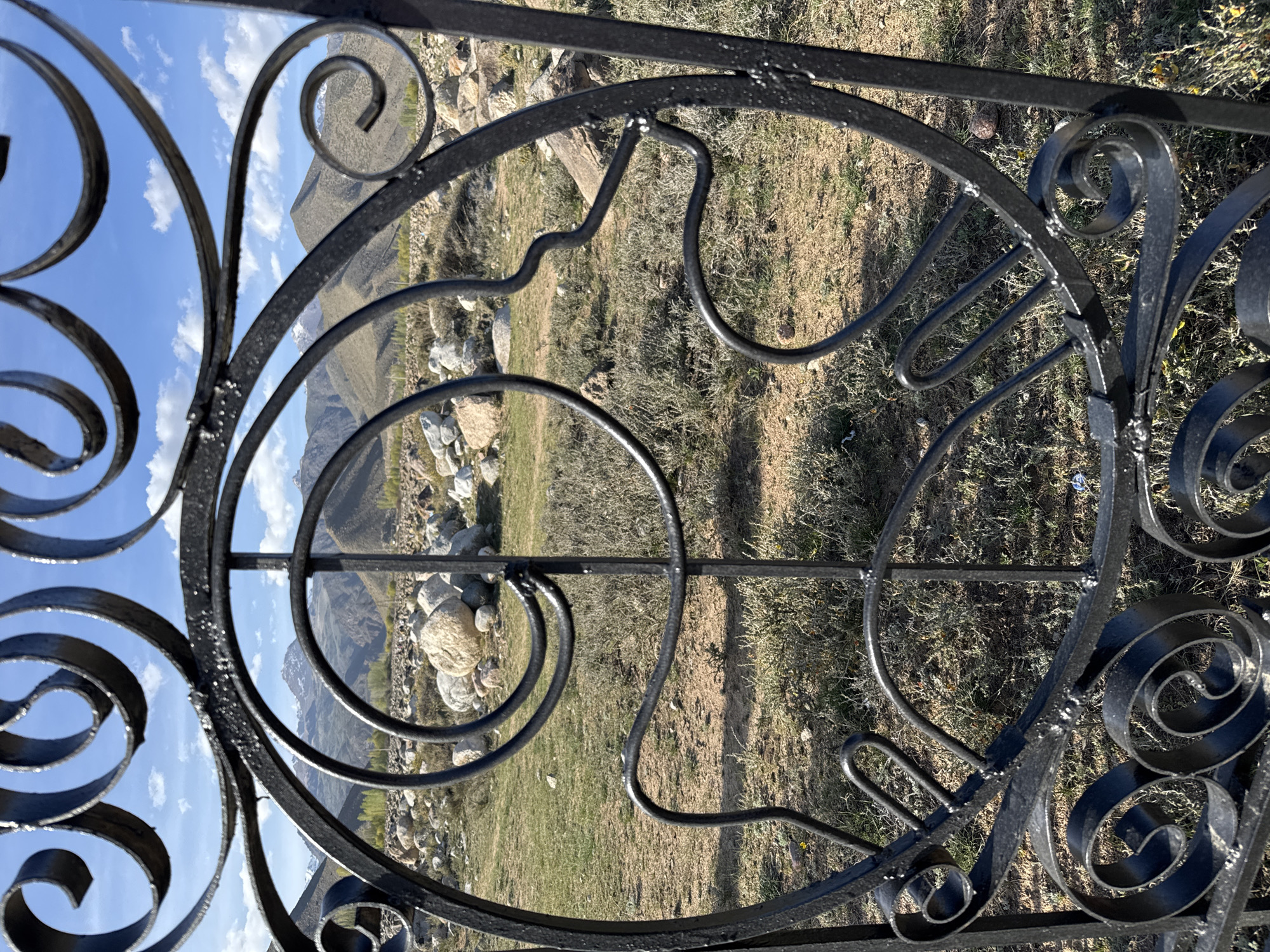 The image shows a decorative black metal gate, likely a fence or entrance feature. The design incorporates curves and swirls. The gate is positioned in front of a landscape featuring grassy terrain, rocks, and mountains in the background. The sky is visible through the gate's design, showing a blue sky with clouds. Based on the context of Çolpon-Ata, Kyrgyzstan, the surrounding environment suggests a mountainous region with natural scenery.