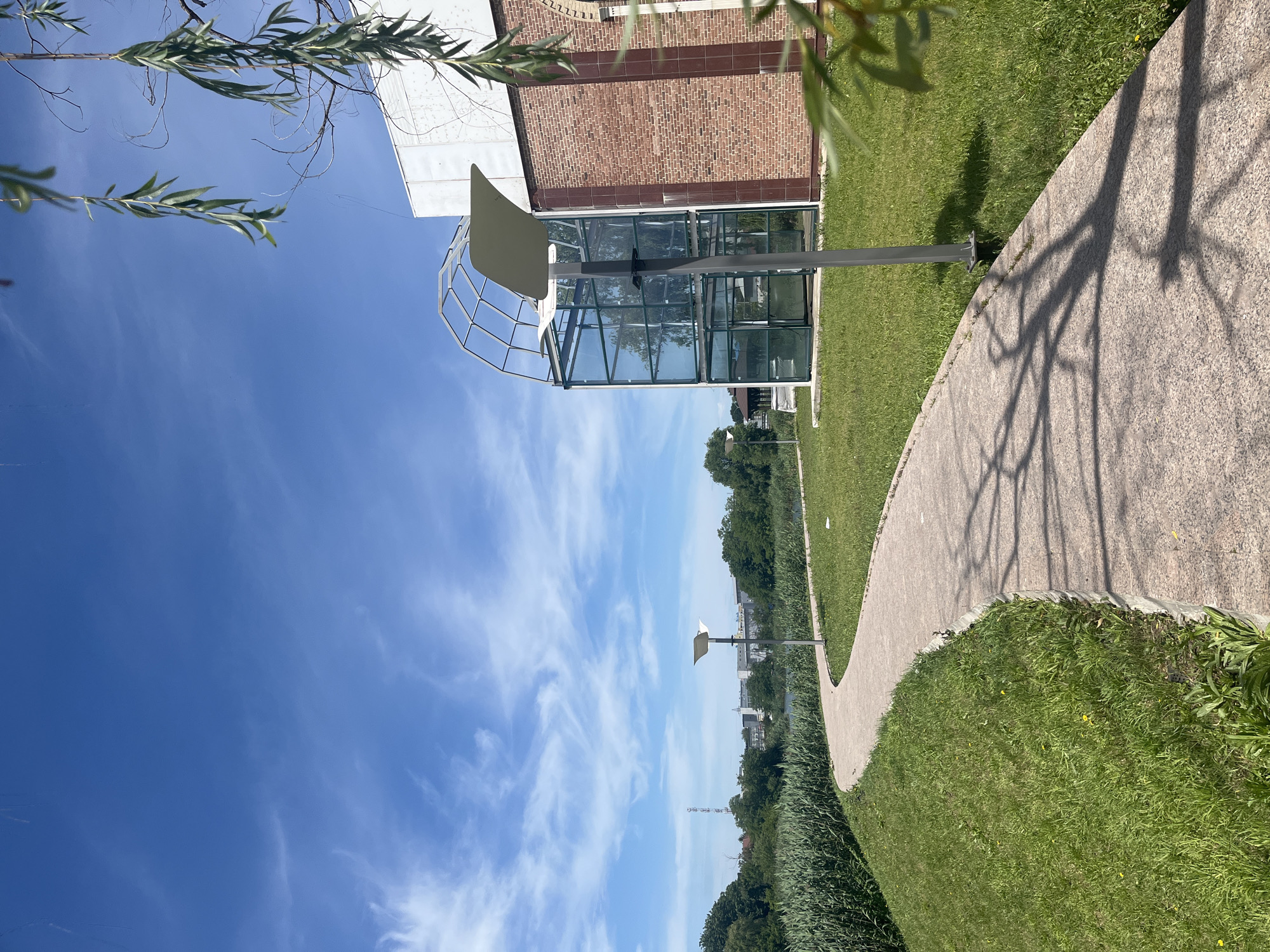 The image displays a bright, sunny outdoor scene in what appears to be a park or promenade area in Krasnodar, Russia. The sky is a clear, vibrant blue, adorned with soft, wispy white clouds.
In the mid-ground, a modern architectural structure stands out. It features a building with a reddish-brown brick facade on its upper section, transitioning into a contemporary glass-paneled lower section. A distinctive, light-colored, curved canopy or roof, supported by a metal framework, extends over this glass part. A modern lamppost with a curved white shade is positioned near this structure.
A light-colored, textured path, possibly concrete or gravel, curves gracefully through lush green grass, leading from the foreground towards the distance. Distinct, dark shadows of tree branches are sharply cast across this path, indicating strong sunlight.
In the upper left quadrant of the frame, the slender, long green leaves of a tree, possibly a willow, extend into view. The foreground is dominated by well-kept green grass. Towards the horizon, a dense band of dark green foliage stretches across the scene, beyond which faint outlines of distant buildings and a tall transmission tower are visible.
The scene captures a peaceful and serene ambiance on a clear day. There are no people visible, and no specific activities are taking place. No text is visible in the image. The overall impression is one of a well-maintained urban or suburban green space.