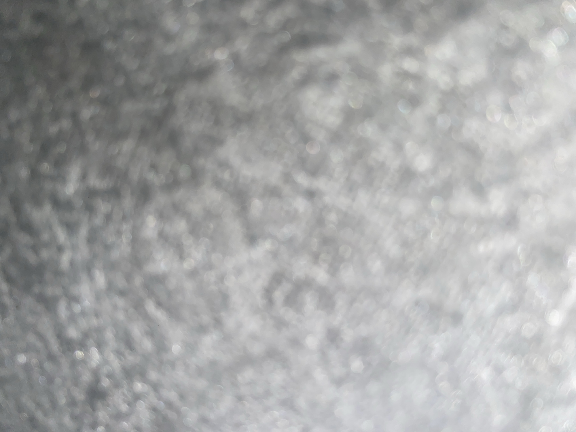 The image displays a blurred, out-of-focus texture that appears to be a metallic or reflective surface. There are numerous small, circular highlights scattered throughout, suggesting a shimmering or glittering effect. The overall color palette is predominantly gray and white, with some subtle hints of iridescent colors within the highlights. The texture seems to be uneven, with variations in light and shadow creating a sense of depth. There are no discernible objects, people, or specific environmental features visible due to the extreme blurriness of the image. The location in Ablanitsa, Bulgaria, cannot be determined from the visual information provided in the image.