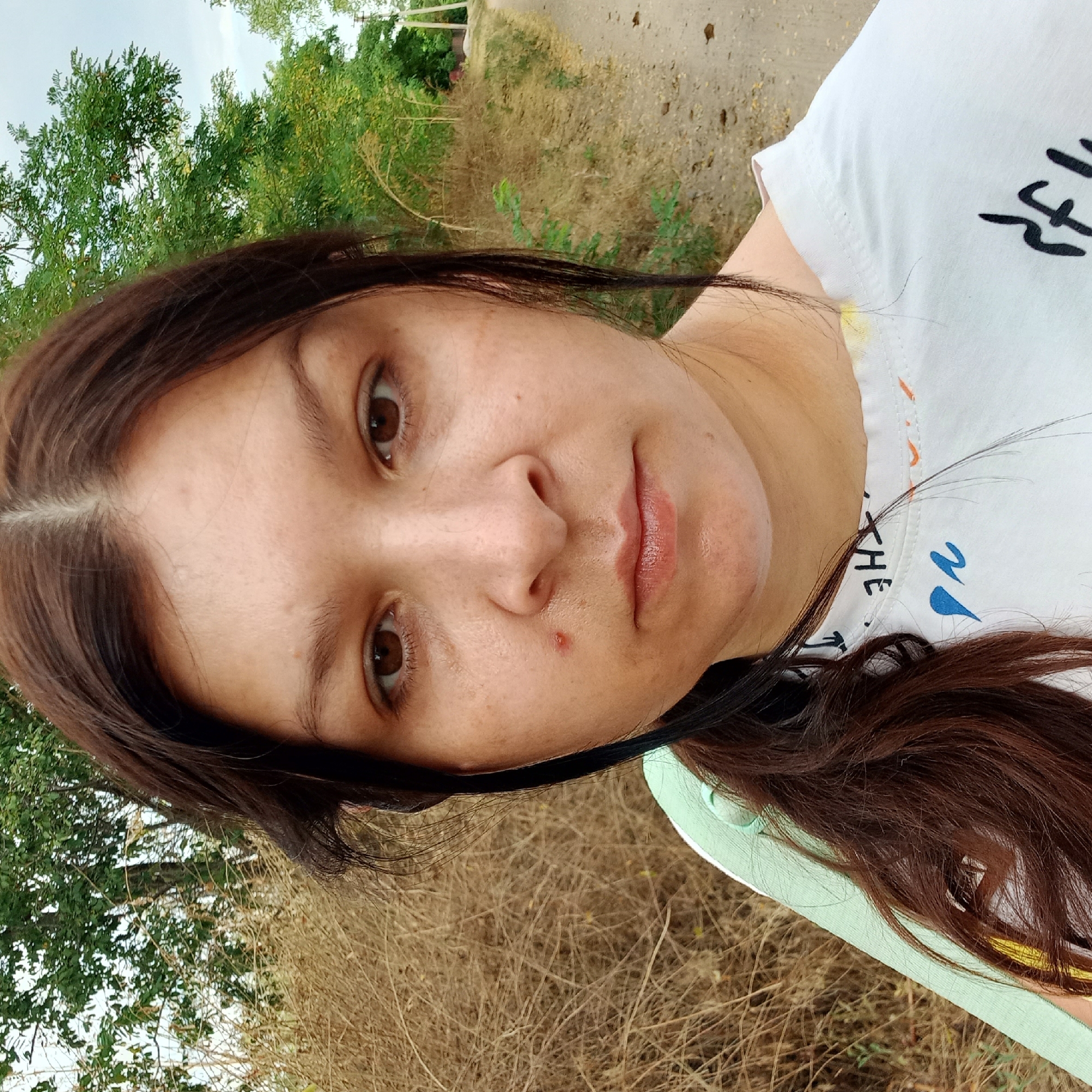 The image is a close-up selfie of a woman with long, dark brown hair. She is wearing a white t-shirt with colorful markings and a light green garment over her left shoulder. Her expression is neutral, and her eyes are looking directly at the camera. A single red pimple is visible on her left cheek. The background appears to be an outdoor setting with dry grass and green trees. A dirt path runs along the right side of the frame. The location is identified as Klyntsi, Ukraine, which is a city in Russia, not Ukraine.