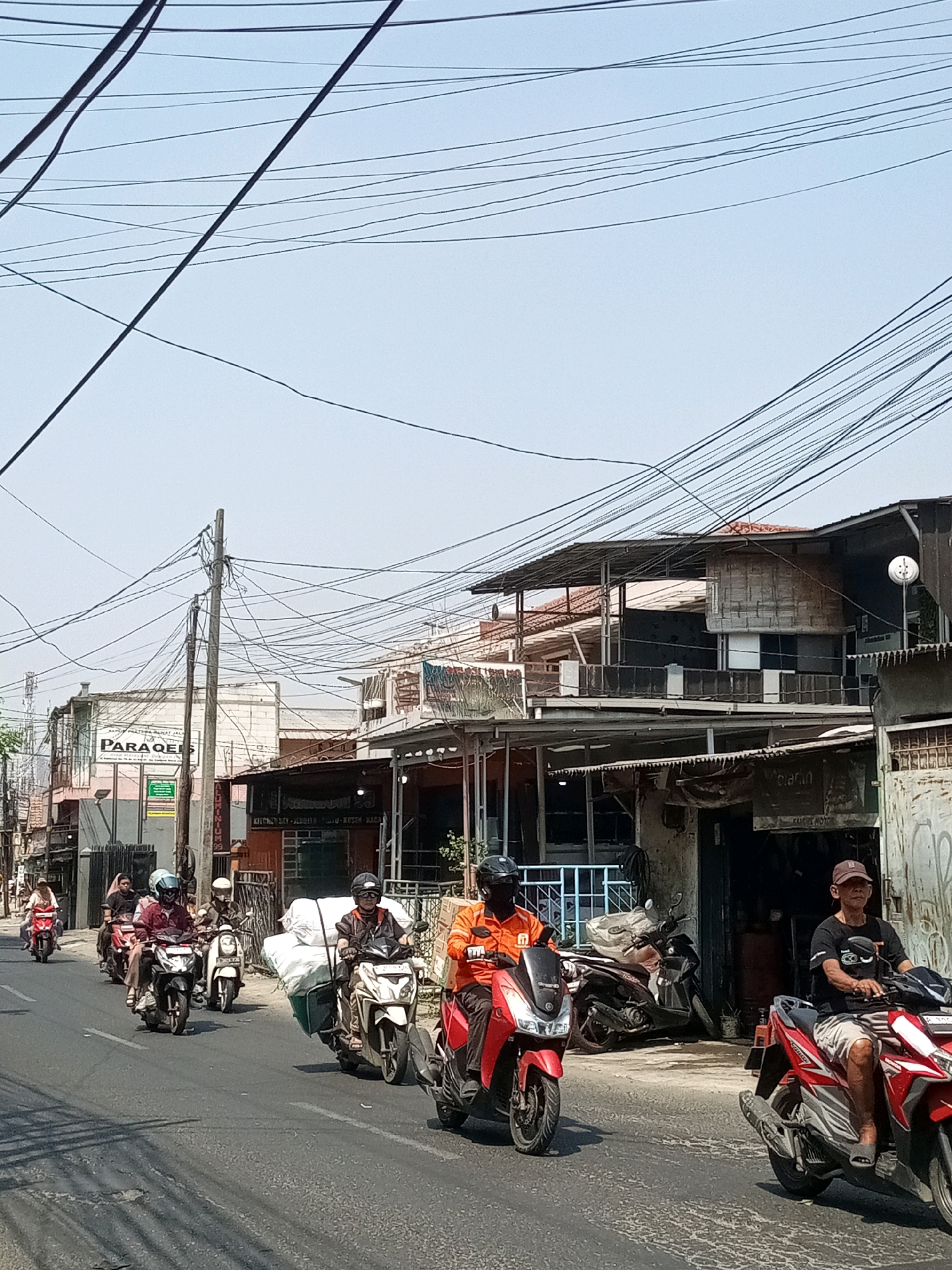 A street scene in Bekasi, Indonesia shows several people riding motorcycles. In the foreground, two men on motorcycles are traveling side-by-side. The man on the right is wearing a black t-shirt, shorts, and a baseball cap, and is riding a red and white motorcycle. The man on the left is wearing an orange shirt and black helmet, and is riding a red motorcycle. Behind them, a line of motorcycles is moving away from the camera. The motorcycles are carrying various items, including large white bundles and what appear to be boxes. The buildings along the street are one to two stories high and appear to be shops or residences. Many power lines are visible crisscrossing the sky above the street. The overall impression is a busy street on a sunny day.