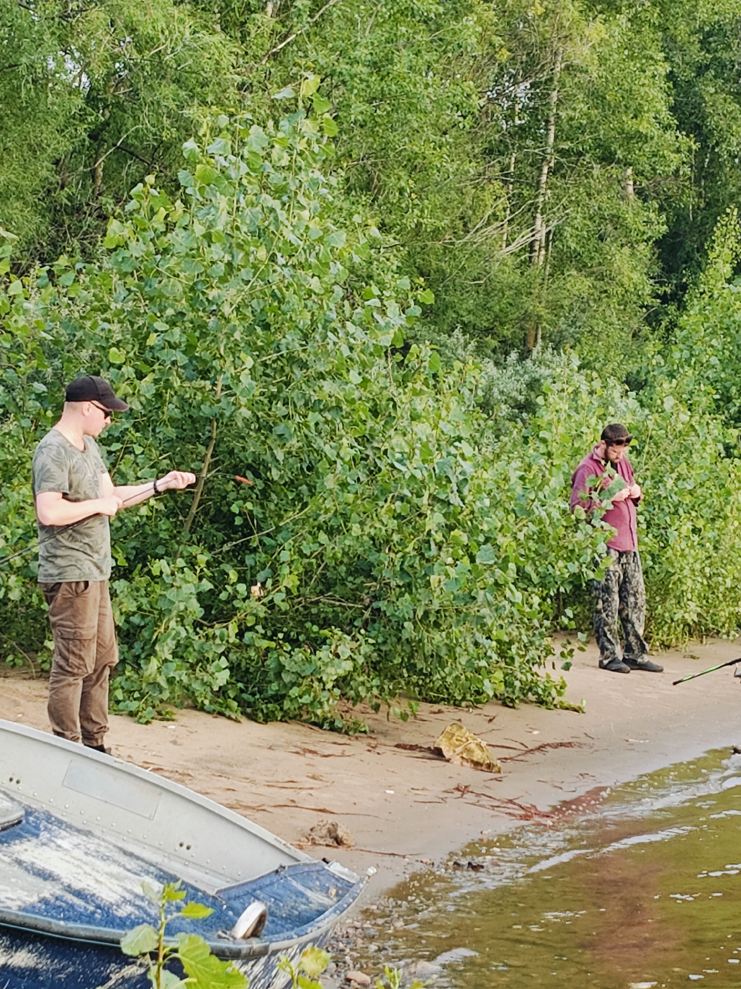 The image shows two men on the shore of the Kama River in Chaikovsky, Russia. The man on the left is wearing a camouflage t-shirt, brown cargo pants, a black baseball cap, and sunglasses. He is holding a fishing rod and appears to be fishing. The man on the right is wearing a maroon long-sleeved shirt and camouflage pants. He is looking down at something in his hands. There is a blue and gray boat on the shore in the foreground. The background is filled with green trees and foliage. The ground is sandy.