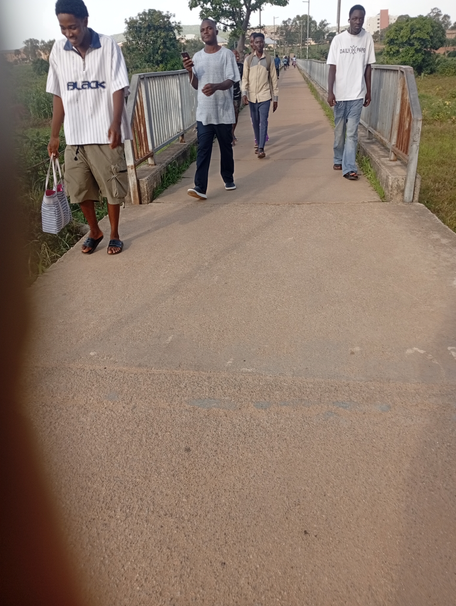 The image shows a group of young men walking on a concrete pedestrian bridge in Rafin Par, Nigeria. The bridge has metal railings on the sides. The men are dressed in casual clothing, with some carrying bags. The environment appears to be semi-urban, with some vegetation visible in the background. The time of day appears to be late afternoon or early evening, given the lighting conditions.