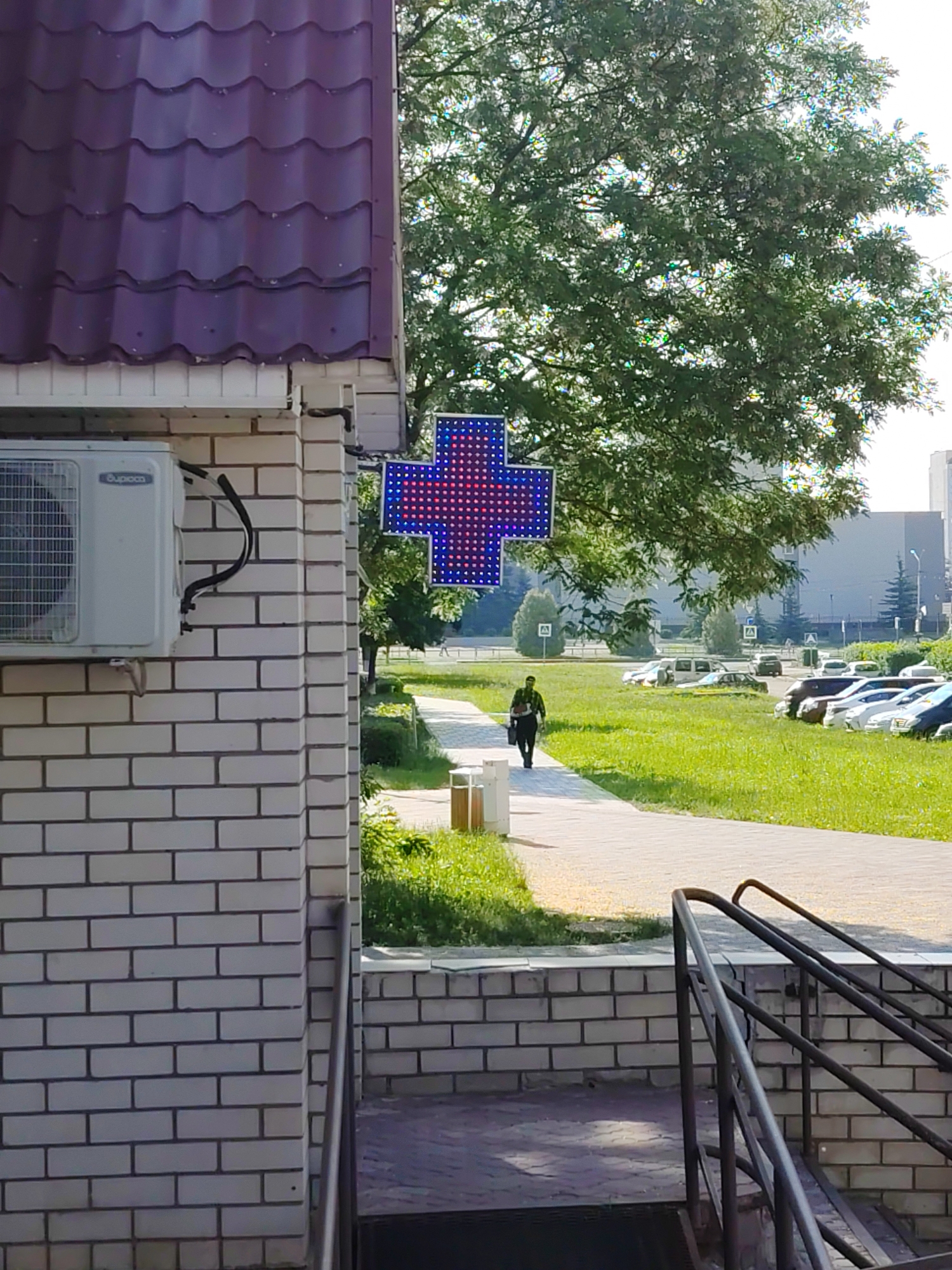 The image depicts an exterior view of a building with a purple tiled roof and white brick walls. An air conditioning unit is mounted on the left side of the building. A large, illuminated blue and red cross, a common symbol for pharmacies, is displayed on the building's facade. In the background, a paved walkway runs through a grassy area where several cars are parked. A lone figure is walking along the path. Trees and buildings form the backdrop of the scene. The context provided by the user indicates this scene is in Desnogorsk, Russia.