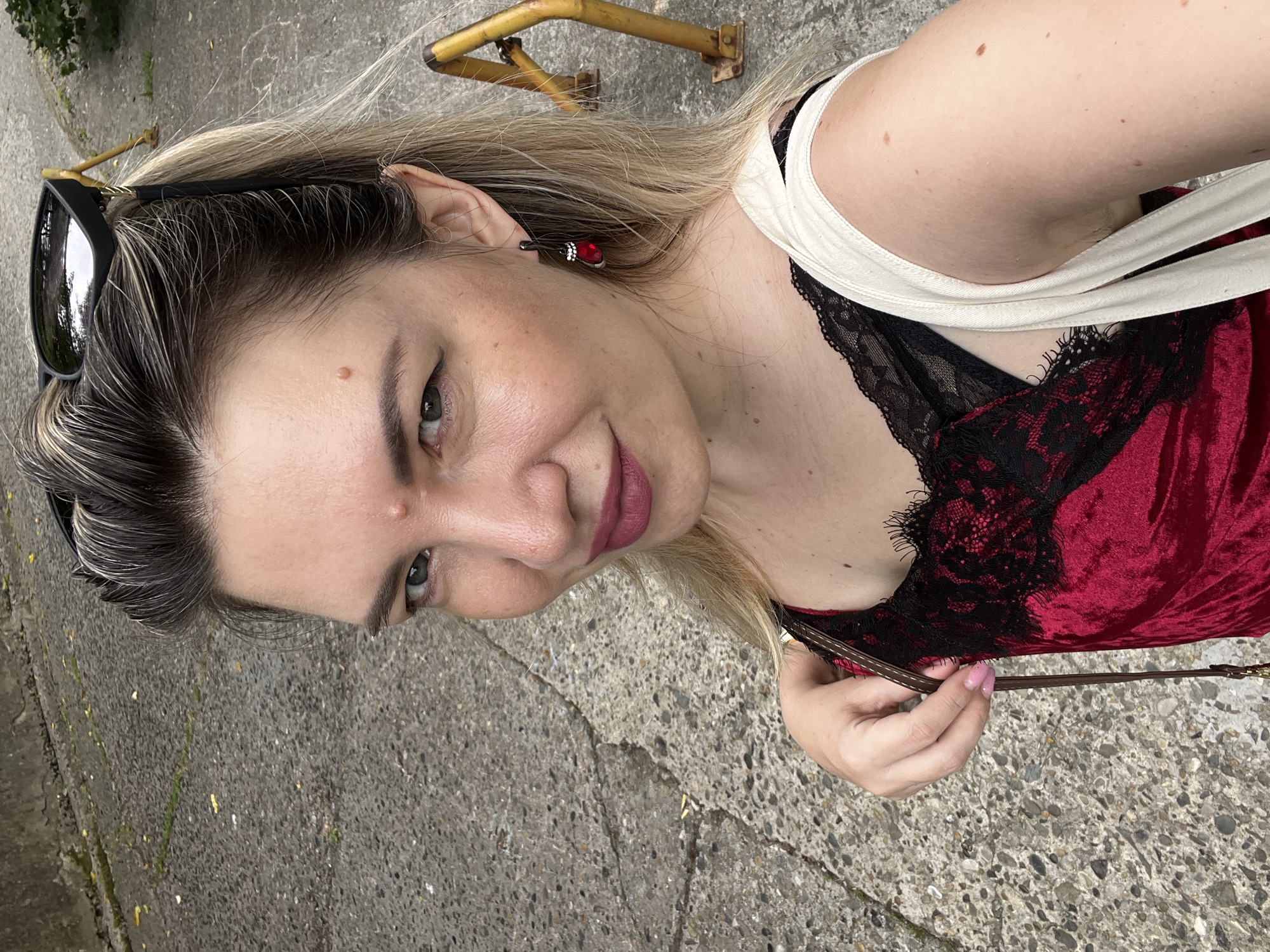 A woman with blonde hair and sunglasses perched on her head is taking a selfie outdoors on a grey, cracked asphalt surface. She is wearing a red velvet dress with black lace trim and a white strap over her shoulder. A red earring dangles from her left ear. Behind her, a yellow metal structure, possibly a bicycle rack or bollard, is visible on the pavement. The background suggests an urban environment, possibly in Sochi, Russia.