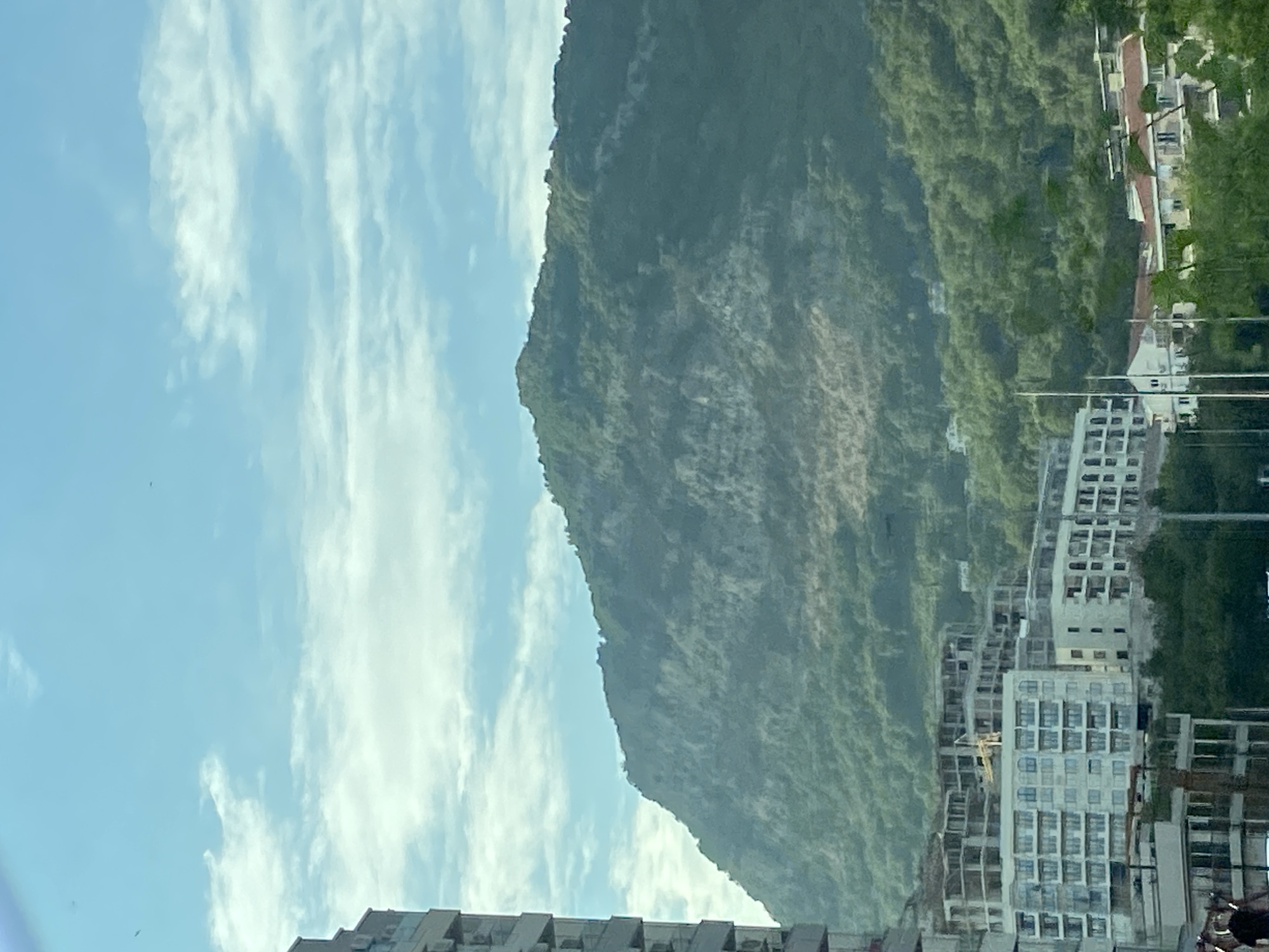 The image shows a view of a coastal city with a mountain range in the background. In the foreground, there are several multi-story buildings, some of which appear to be under construction with exposed frameworks and scaffolding, indicative of ongoing development. One prominent building in the mid-ground is a light-colored, multi-story structure with many windows. To the left, a portion of a tall, modern building with visible air conditioning units is cut off by the edge of the frame. The mountain range behind the city is covered in dense green vegetation, with some lighter patches suggesting rocky outcrops or cleared areas. The sky is blue with scattered white clouds. The scene is consistent with a developing urban area in Budva, Montenegro, a coastal city known for its mountainous backdrop and construction projects.