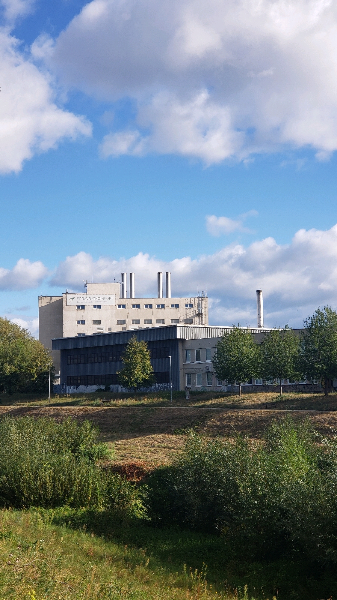 The image shows a multi-story industrial building with several large ventilation pipes on its roof. The building is light-colored and appears to be made of concrete. There are multiple windows on each level. A sign on the building reads "SPRAVBYTKOMFORT". In front of the main building is a darker, lower section with more windows. The building is situated in an area with trees and grass. The sky above is blue with scattered white clouds. The scene is a clear, daytime shot of an industrial facility in the city of Prešov, Slovakia.