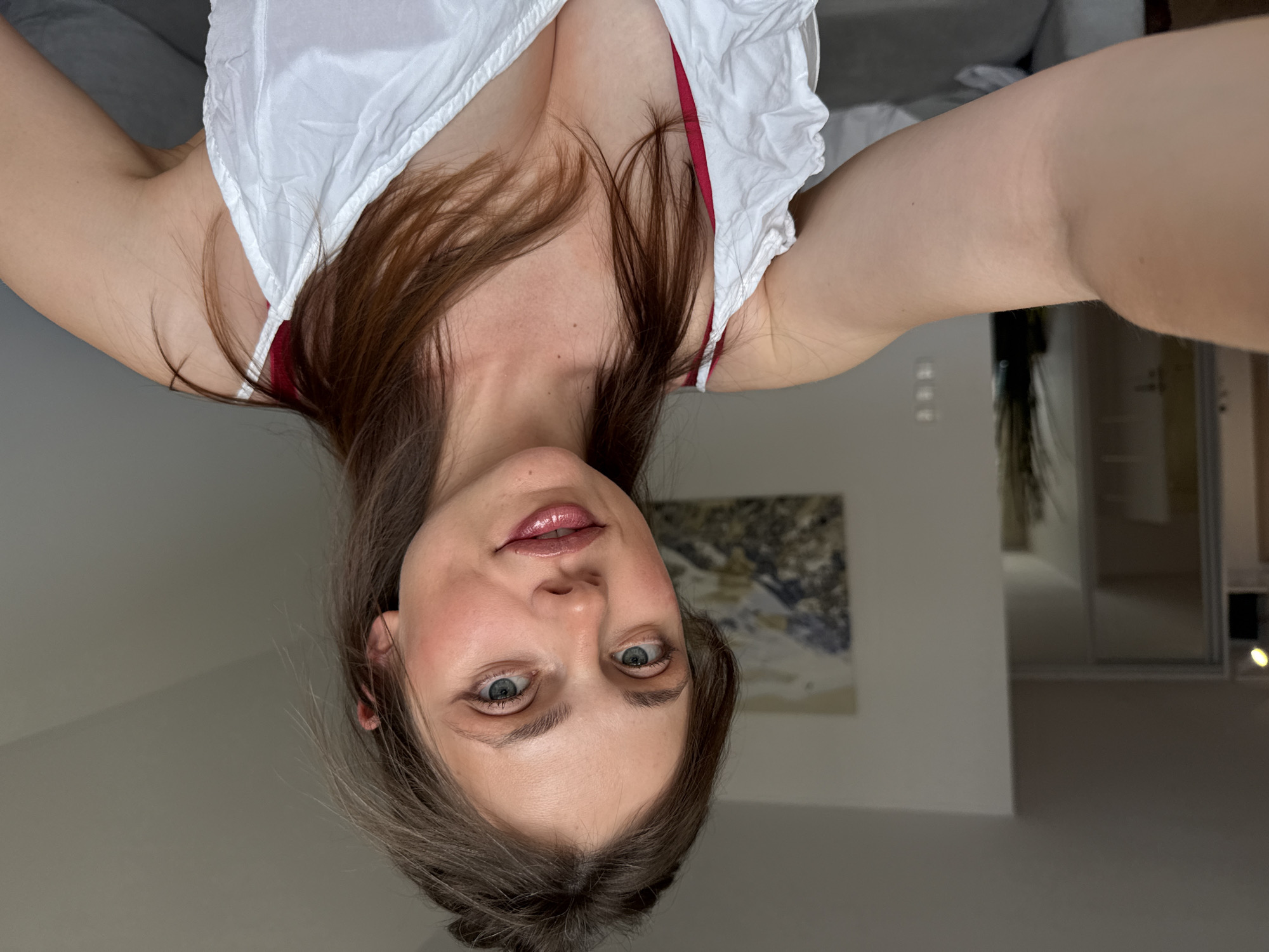 The image is a selfie taken by a young woman with light brown hair, blue eyes, and light skin. She is wearing a white top with red straps, and she has makeup on, including eyeliner and lipstick. The woman is indoors, and the background appears to be a home interior. There is a couch and a painting on the wall visible in the background, as well as a large closet with sliding doors. The lighting suggests it is daytime. The woman is looking directly at the camera, and she is smiling slightly. She is holding the camera with her right hand, which is partially visible in the frame. The image does not depict any specific landmarks or elements related to Szklarska Poręba, Poland.