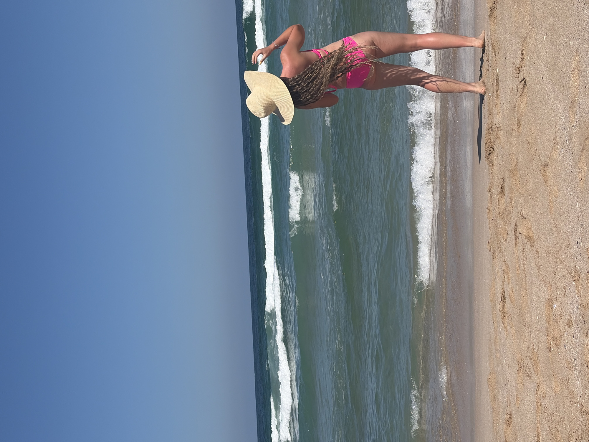 A person wearing a pink bikini and a wide-brimmed straw hat stands on a sandy beach, facing the ocean. Their hair is long and braided, extending down their back. The person appears to be holding their arm up, possibly adjusting the hat or taking a photo. The ocean waves are breaking on the shore, and the sky is clear and blue. The scene is likely on a sunny day at a beach.