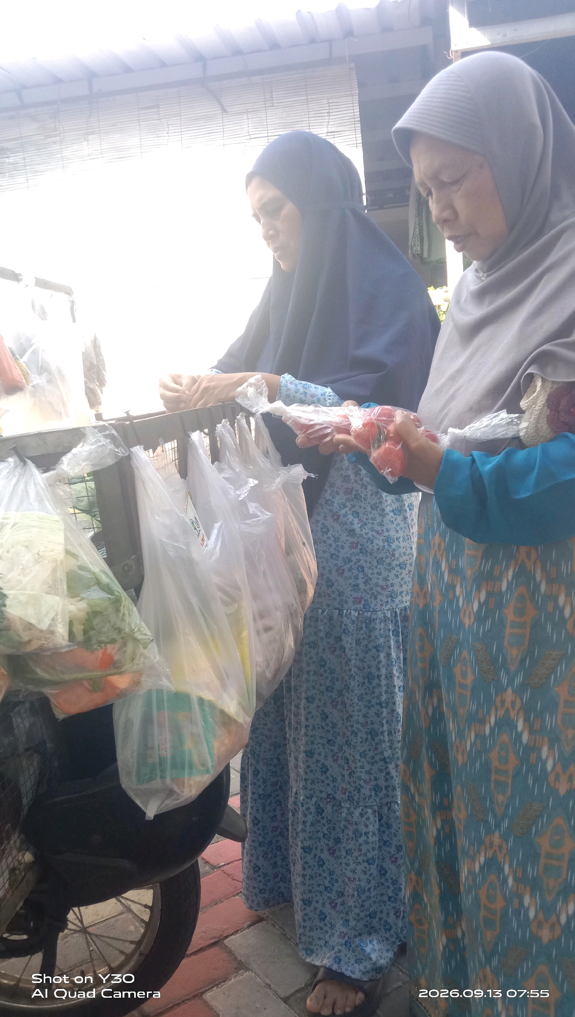 Two women are standing next to a food cart in Sekarkurung, Indonesia. The woman on the left wears a dark blue hijab and a blue floral dress. The woman on the right wears a grey hijab and a blue and orange patterned dress. Both women appear to be interacting with the food cart, which is laden with plastic bags filled with produce. The woman on the right is holding a bunch of small red tomatoes wrapped in plastic. The background shows a woven blind and a roof structure. The ground is paved with brick. The timestamp "2026.09.13 07:55" is visible in the lower right corner.