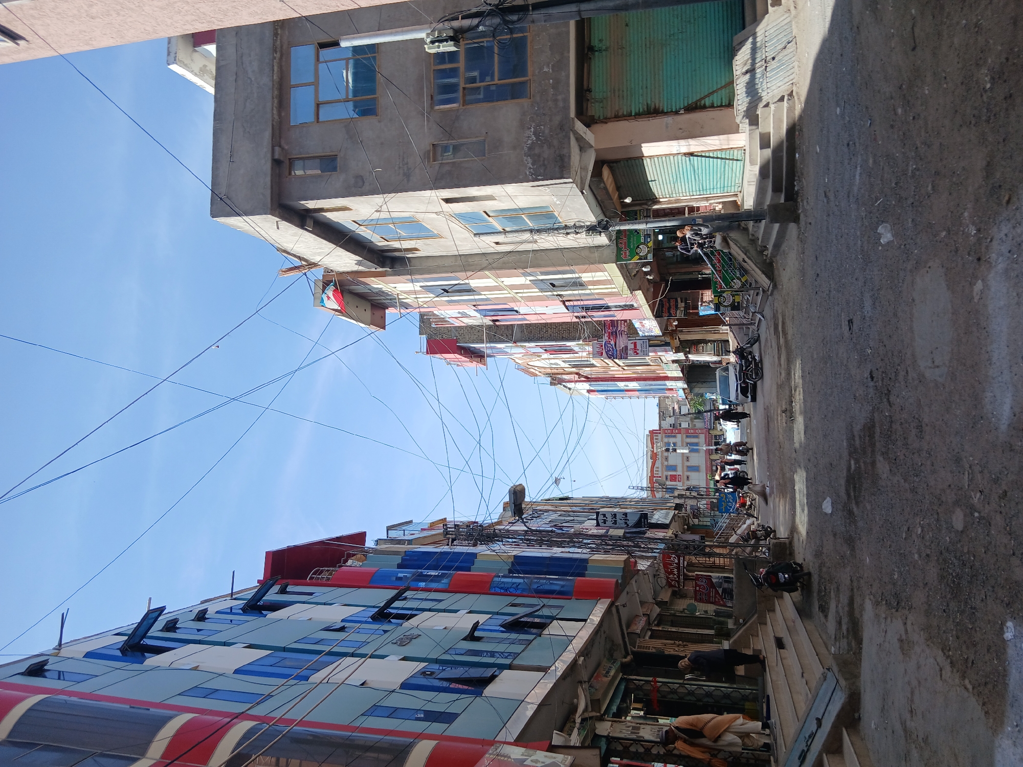 The image captures a vibrant street scene in Quetta City Tehsil, Pakistan, under a clear blue sky. The photo is taken looking down a narrow, dusty street lined with multi-story commercial buildings on both sides.
On the left, a prominent building features a modern facade with blue, red, and grey panels and windows that appear to be partially open, resembling awnings. Other buildings along this side are made of concrete with various window styles. The sky above is filled with a dense network of crisscrossing power lines and cables, a common sight in urban areas of the region.
On the right side of the street, more concrete buildings house shops, many with roll-down metal shutters, some painted green. Steps lead up to several of these storefronts. A few individuals are visible on the street and near the shops, including one person in what appears to be traditional attire. Several motorcycles are parked along the right side of the road, and a white car is partially visible further down the street.
The scene is brightly lit by strong sunlight, casting sharp shadows on the street and buildings, suggesting it is midday or early afternoon. Shop signs in Urdu script are visible, with prominent examples including "ہربل" (herbal) on a green sign and "کریَم" (cream) on another, indicating businesses like pharmacies or general stores. The overall impression is that of a busy, functional urban commercial district.