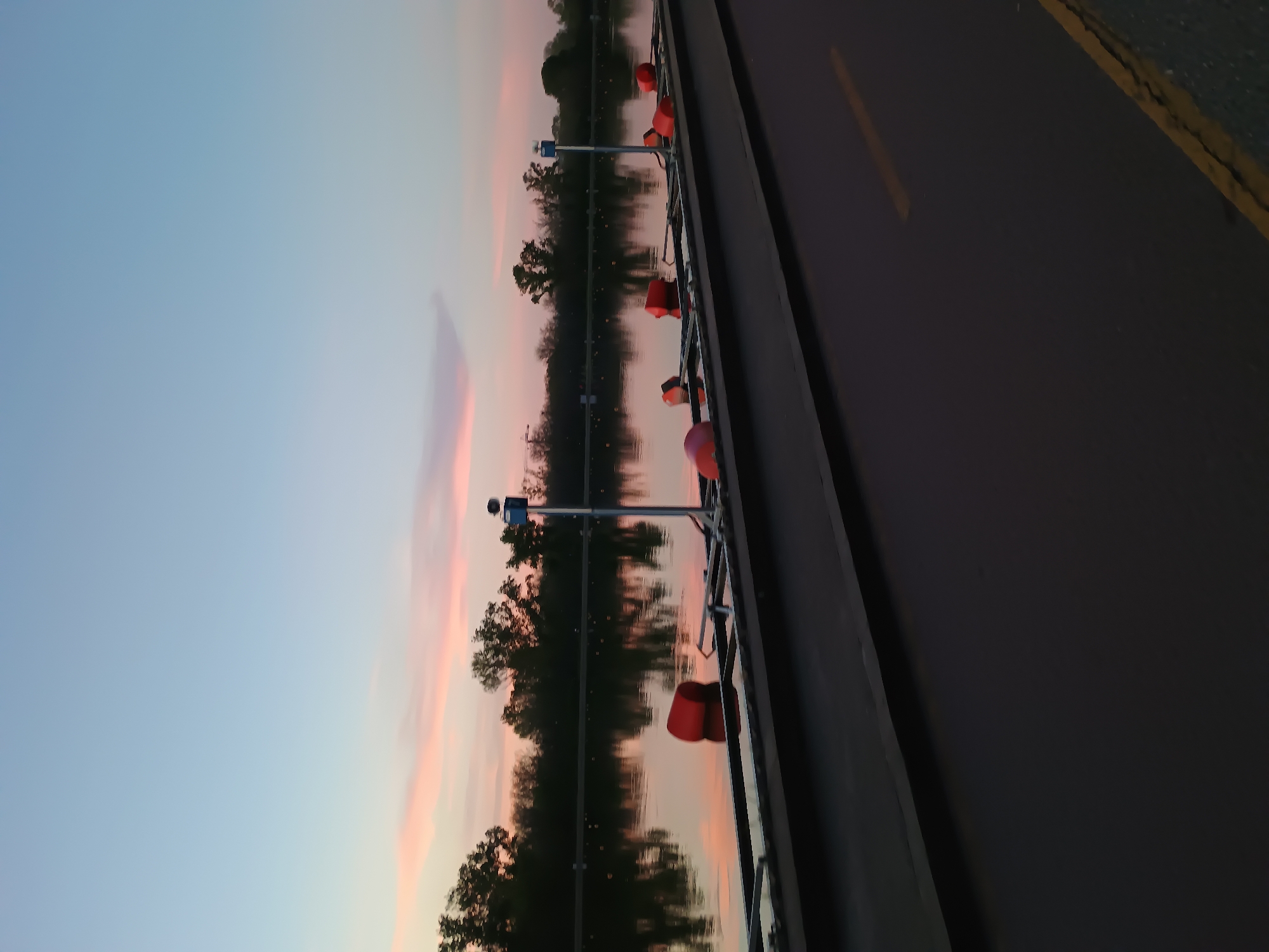 The image depicts a scene at sunset, likely in Plovdiv, Bulgaria, based on the presence of a water channel and elements consistent with the city's landscape. The sky is a blend of light blue and pink hues, reflecting the setting sun. A body of water, possibly the Maritsa River or a canal associated with it, mirrors the sky and the silhouetted trees lining its banks. Along the edge of the water, there are evenly spaced metal poles with red, buoy-like objects attached. To the right of the water is a gray road surface with a single yellow line marking the edge of the road. The image is oriented sideways, with the sky and water on the left and the road on the right. The lighting is dim, suggesting the onset of twilight.