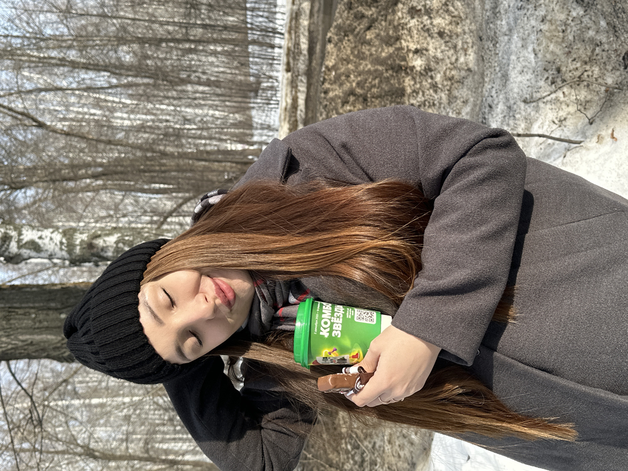 A young adult female is depicted outdoors in a sunlit, wooded area within Chaikovsky, Russia, during what appears to be late winter or early spring. She wears a black knitted beanie, a grey patterned scarf, and a dark grey winter coat. Her eyes are closed, and her lips are pursed, with her head tilted slightly. Her right hand is positioned near the back of her head, touching her hair or hat. In her left hand, she holds a green disposable cup with Cyrillic text "КОМБО ЗВЁЗД" and a QR code, along with a partially unwrapped chocolate bar. The background consists of a dense arrangement of bare deciduous trees, including several birch trees, with patches of melting snow and exposed earth on the ground.