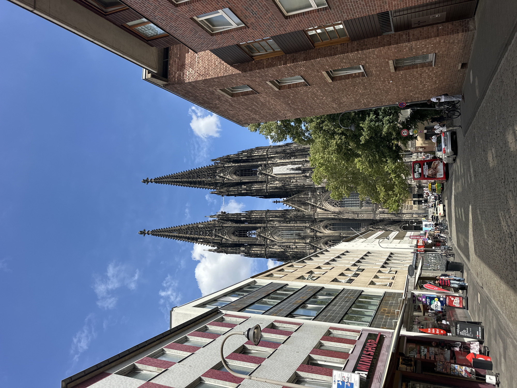 The image depicts a street scene in Cologne, Germany, with the Cologne Cathedral prominently visible in the background. The cathedral is a large Gothic structure with two tall spires reaching towards a blue sky with scattered white clouds. The street is paved with cobblestones and lined with buildings on both sides.
On the left side of the street, there are several multi-story buildings. The closest building has a facade with alternating horizontal bands of light-colored and red-striped material. A street lamp with a curved arm is attached to this building. Further down the street on the left are more buildings with windows and storefronts, including one labeled "MINI SHOP."
On the right side of the street, there are buildings made of red brick. These buildings have windows and some have darker vertical siding elements.
The street is active with people and vehicles. Several people are walking on the sidewalks and crossing the street. A silver car is parked on the right side, and a red double-decker bus, labeled "CityTour," is driving down the street. Bicycles are also visible, parked on the sidewalk on the right. The overall impression is a sunny day in a bustling urban environment, with the historic cathedral serving as a dominant landmark.