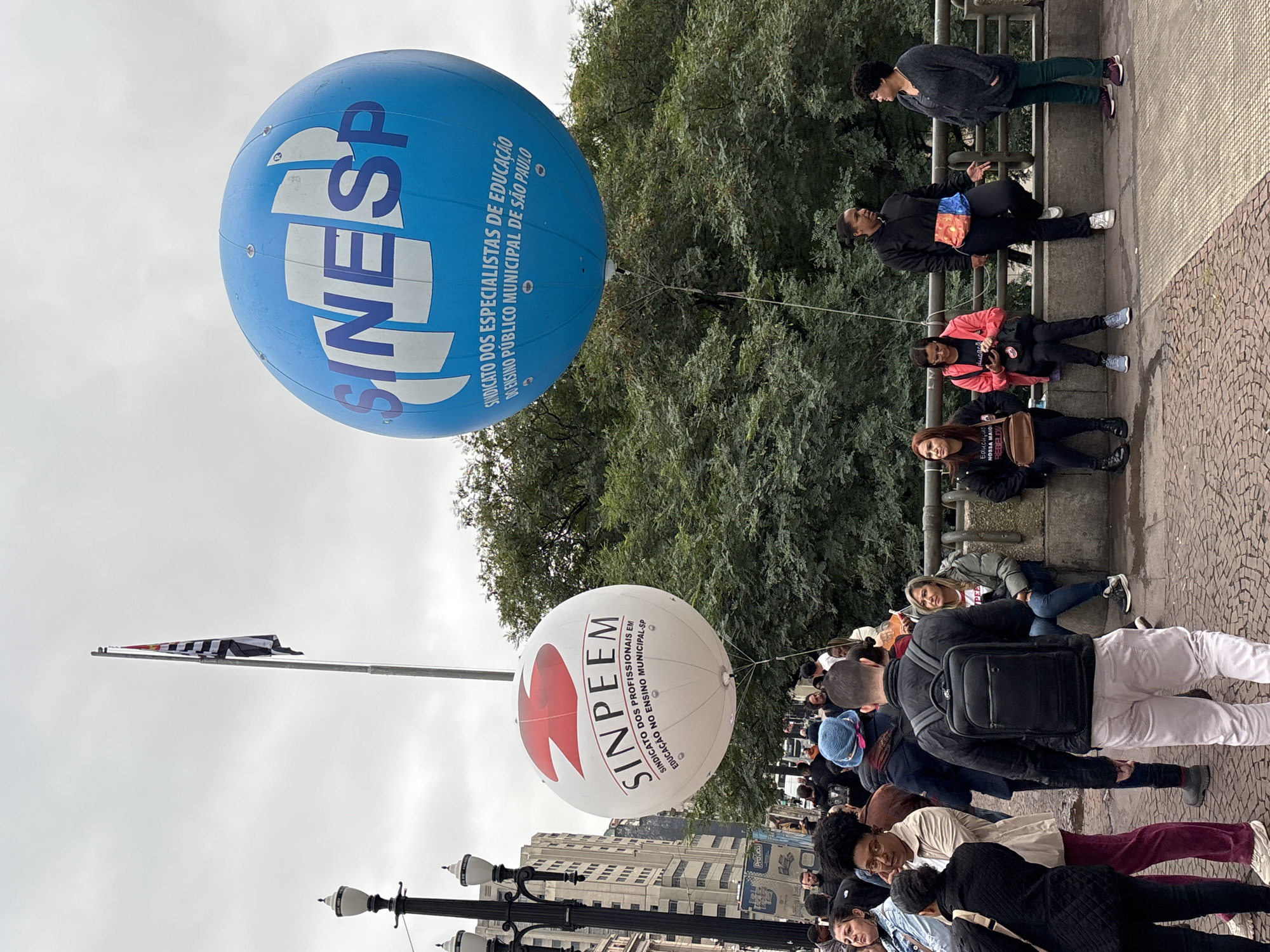 The image shows a street scene in São Paulo, Brazil, with two large, spherical balloons tethered above a crowd of people. The upper balloon is blue with the white text "SINESP" and additional smaller text detailing its association with education specialists in São Paulo's public school system. The lower balloon is white with a large red logo and the text "SINPEEM," also related to education professionals in the municipal public school system.
A crowd of people is visible, primarily standing, with some individuals sitting on a ledge or wall in the background. A flag is seen in the background as well. The sky is overcast. The architecture visible in the background suggests an urban environment. The street is paved and has some decorative elements. The people appear to be at a demonstration or public event.