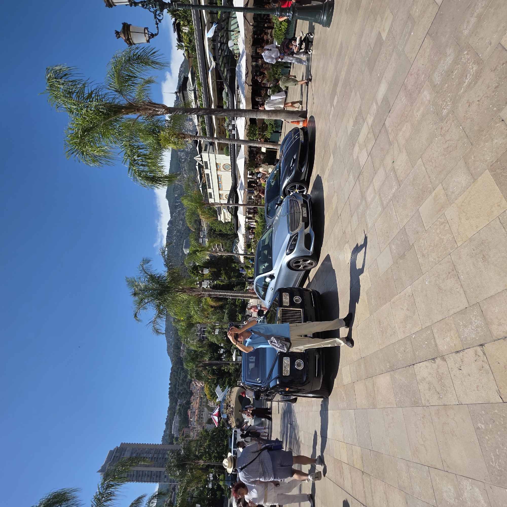A vertical, full shot shows a sunny day in Monaco. The scene is a street paved with light-colored square tiles. On the left, a line of luxury cars is parked. The closest car is a black Rolls-Royce, followed by a silver Mercedes-Benz, and a dark gray coupe. Behind the cars are palm trees and buildings, including a white building with awnings that appears to be a cafe. In the background, a lush green hillside rises, dotted with buildings.
Several people are visible. In the foreground, a man in a light blue polo shirt and beige pants stands by the Rolls-Royce, holding a phone up as if taking a photo. He has a bag slung over his shoulder. To the left and slightly further back, another man in a plaid shirt and shorts, wearing a straw hat, walks away from the camera. A woman in a white top and light-colored skirt is walking beside him. Further back, more people are seen walking and sitting at outdoor tables in front of the cafe.
The time of day is clearly daytime, indicated by the bright sunlight and long shadows cast by the people and cars. The sky is a clear, vibrant blue, suggesting good weather. The presence of palm trees and the architecture of the buildings, as well as the luxury cars, are strong cues pointing to Monaco as the location. The overall atmosphere appears relaxed and leisurely, with people enjoying the sunshine and the picturesque surroundings. There is no visible text other than a license plate on the Mercedes-Benz, which reads "MC 67745".