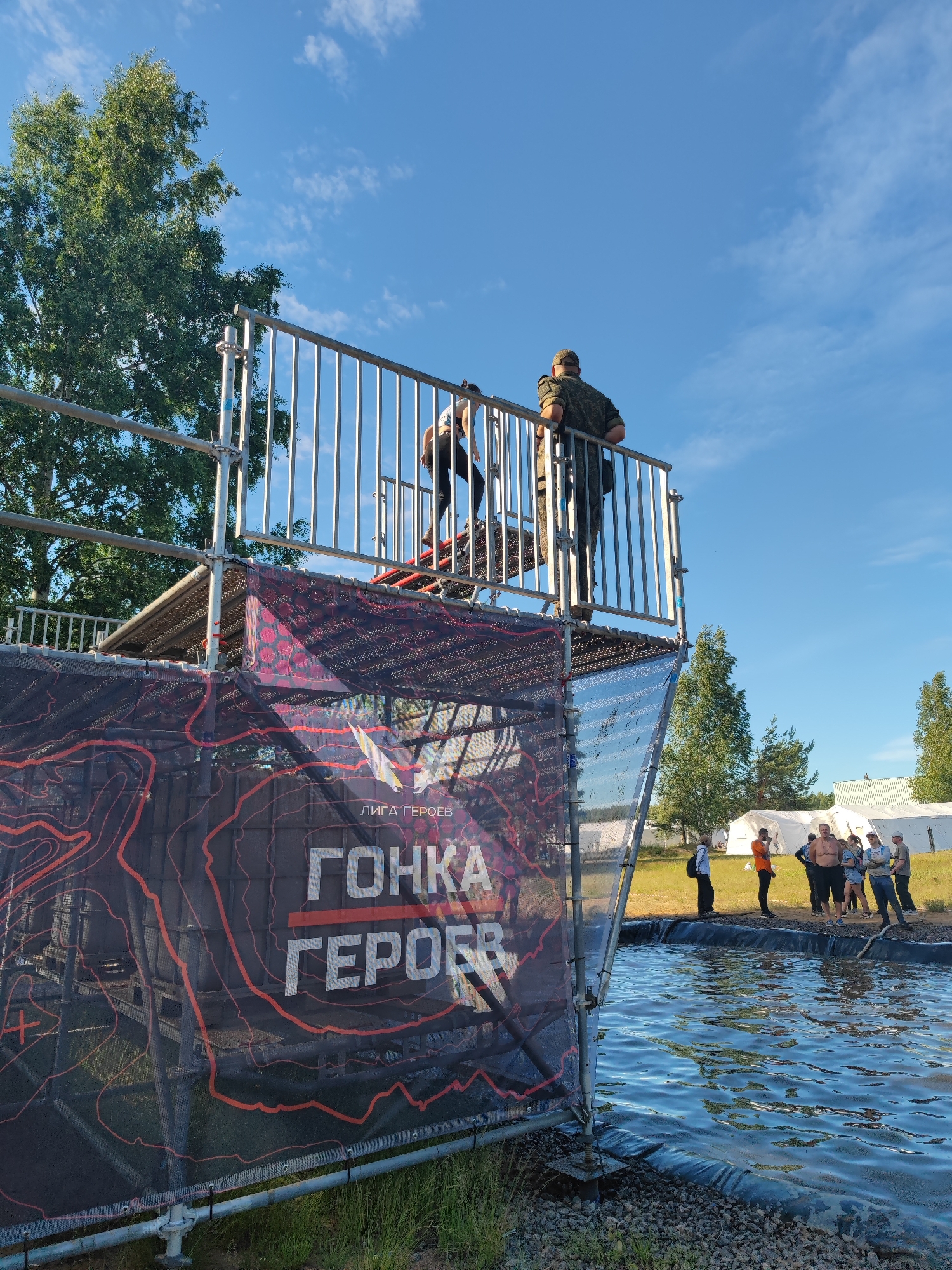 An outdoor scene features a temporary structure, likely part of an obstacle course, positioned adjacent to a body of water. The structure is constructed from metal scaffolding and includes a platform with a railing, from which two individuals are visible. One person, dressed in camouflage attire, stands facing away from the camera, leaning on the railing. Another individual, wearing a white shirt and dark pants, is partially visible on the platform, descending a set of stairs.
A large banner is draped across the front of the structure, displaying the Russian text "ЛИГА ГЕРОЕВ" and "ГОНКА ГЕРОЕВ," which translates to "League of Heroes" and "Race of Heroes." The banner has a dark background with red topographic-like patterns.
In the background, several people are gathered near white tents, suggesting an event or competition is taking place. The environment is grassy with scattered trees, and the sky is clear blue with scattered clouds.
The scene is located at the Sertolovo Sports Complex in Sertolovo, Russia, a venue known for hosting the "Race of Heroes" obstacle course events.