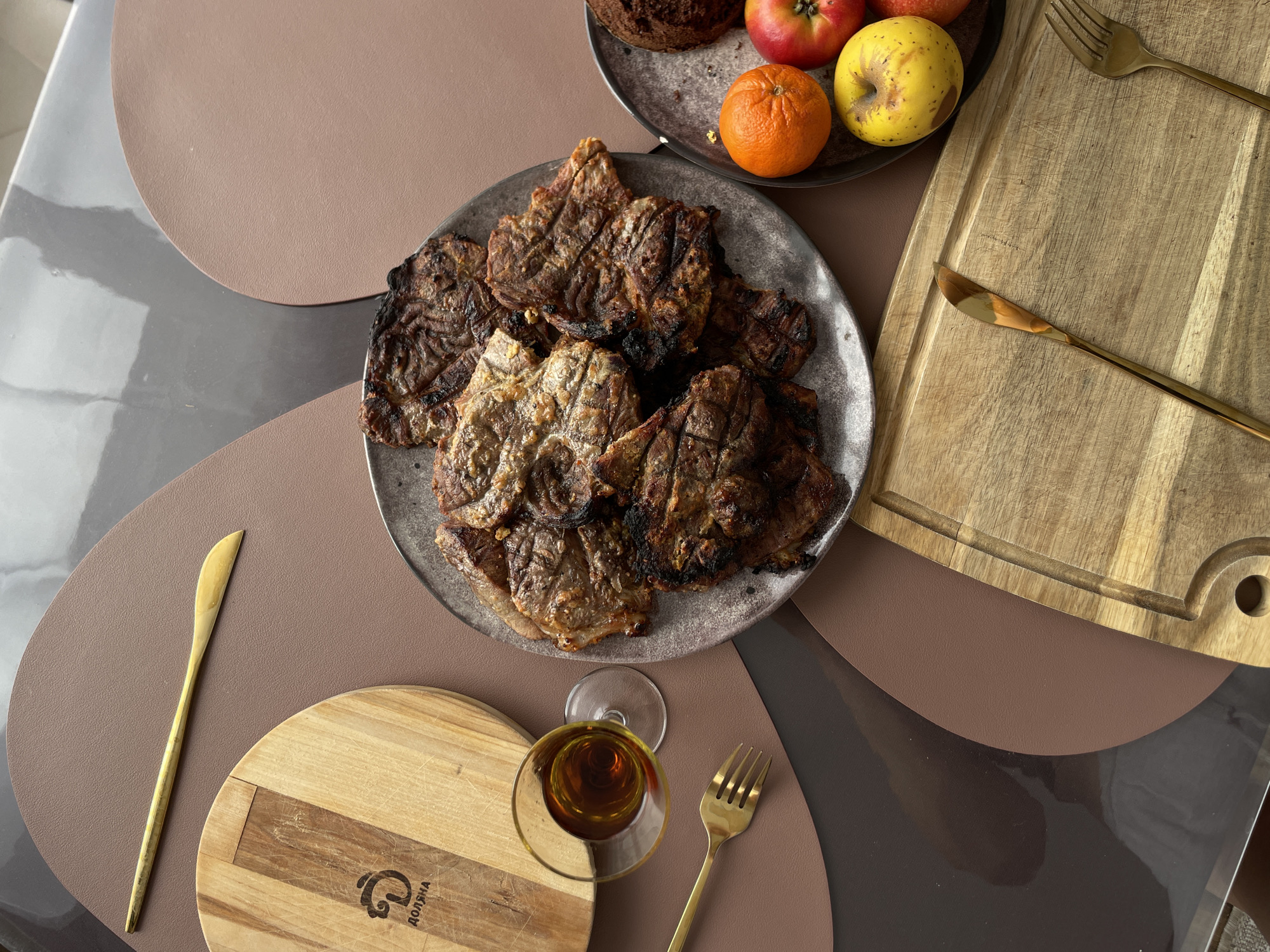 A high-angle shot shows a table setting with a plate of grilled meat as the central focus. To the upper right, another plate contains an apple, orange and muffin. A glass of amber liquid sits near the lower center. A cutting board, fork, and knife are also arranged around the plates and glass. The table's surface is covered with geometric-patterned placemats in brown, gray, and beige tones. The scene suggests a meal setup, possibly in Подшивалово, Russia, given the prompt.