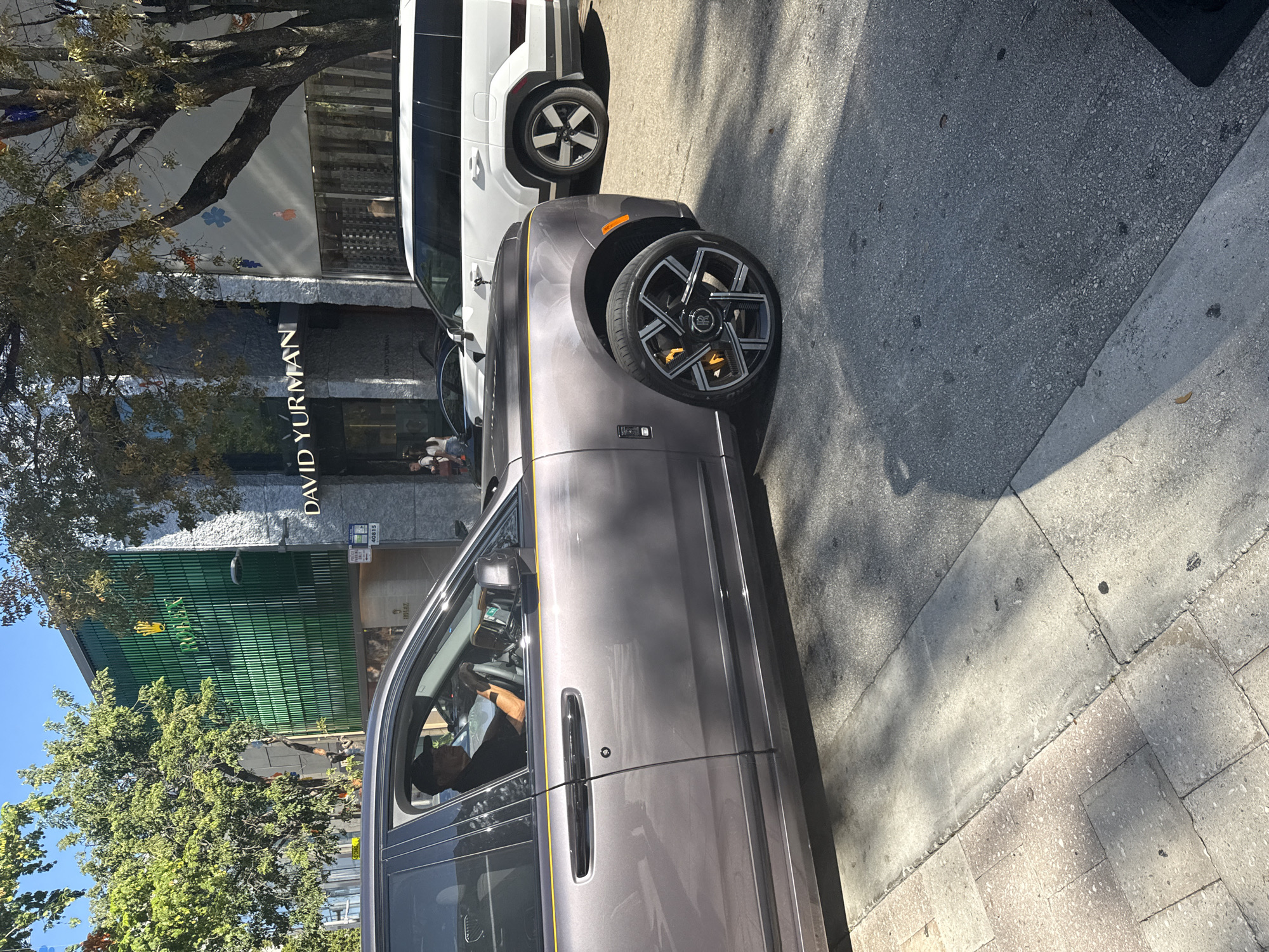 The image captures a street scene in Miami, United States of America. A gray Rolls-Royce and a white Rivian SUV are parked on the side of the road. The storefronts of David Yurman and Rolex are visible in the background. Trees line the sidewalk, casting shadows on the street and buildings. A person is seated inside the Rolls-Royce.