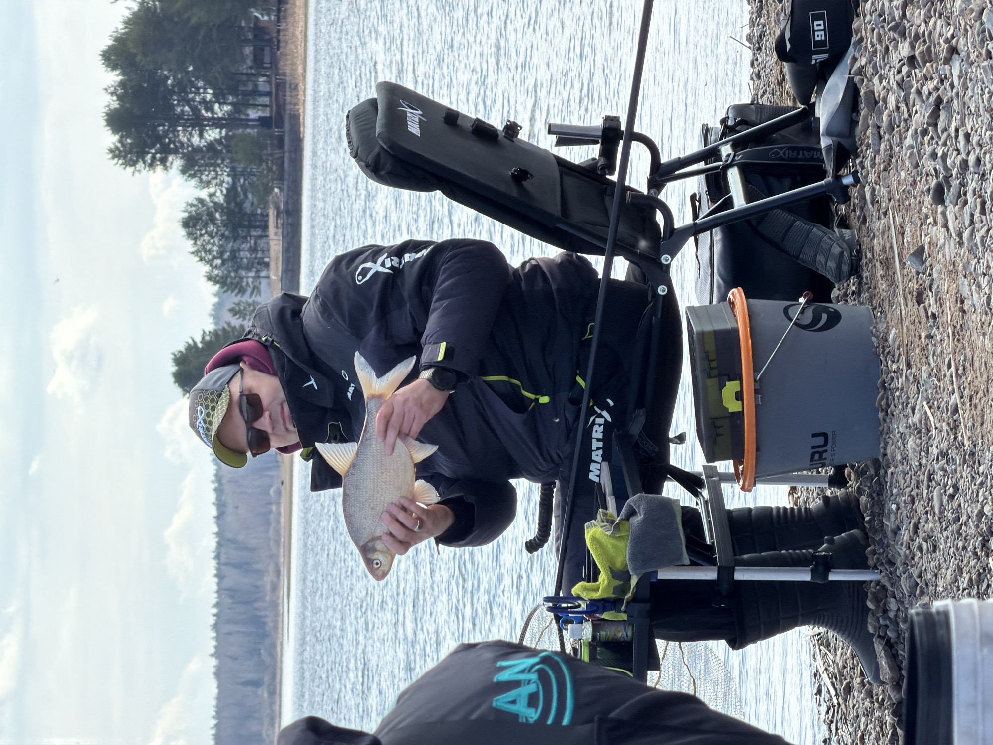 The image, taken in Chaykovsky, Russia, depicts an outdoor fishing scene by a body of water, likely a lake or wide river, under an overcast sky. In the center, a person is seated on a specialized fishing station or chair, holding a medium-sized, light-colored fish, possibly a bream or similar species. The person wears a dark jacket, a baseball cap with a pattern, and sunglasses. A watch is visible on their left wrist.
The fishing setup includes a "MATRIX" branded chair with various attachments. To the right of the person, a grey bucket with orange accents is visible, bearing the text "GURU FEEDER & POWER." Part of a fishing rod extends upwards. In the immediate foreground, the back of another person's jacket is visible, featuring an "AN" logo in light blue. The person holding the fish also has a fish icon and some text on their jacket sleeve. Another piece of equipment near the chair shows "Xanow" branding, and a bag in the background has "90" printed on it.
The setting is a rocky or gravelly shoreline, with the water stretching out behind the subject. The opposite bank features a dense line of pine trees under the cloudy sky. The overall impression is of a quiet, focused fishing outing. The light suggests it is daytime, possibly late morning or afternoon. The person appears to be presenting their catch.
