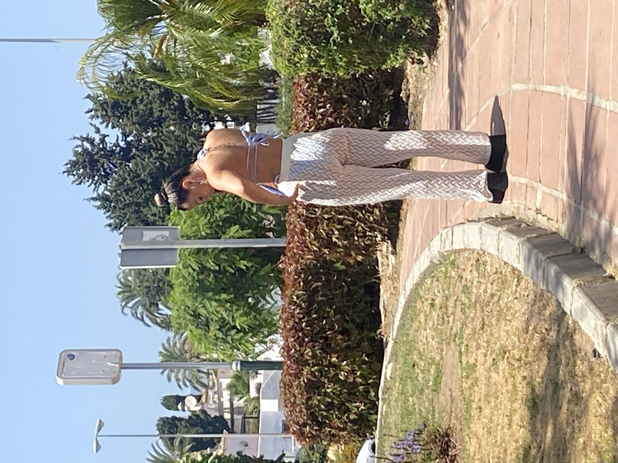 A woman wearing a light blue halter top and white patterned pants stands on a paved walkway in a park on a sunny day. She is facing away from the camera, looking down and adjusting the back of her top. Behind her, a red-leafed hedge forms a border with a grassy area to her left. In the background, there are palm trees, other greenery, buildings, and two street signs on poles. The walkway is made of red-toned rectangular tiles, and a concrete curb separates it from the grass. The scene appears to be located in Estepona, Spain, given the vegetation and architectural style.