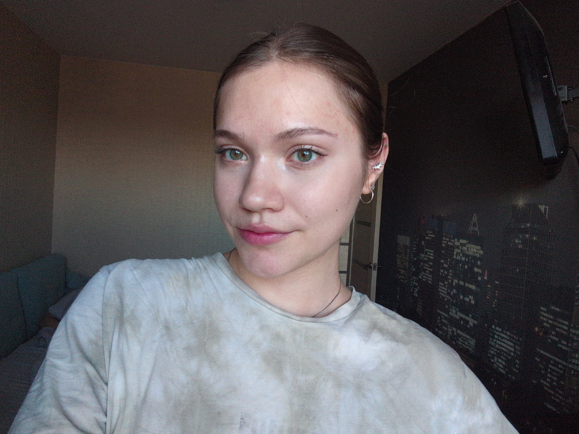 A fair-skinned woman with brown hair pulled back is centered in the frame, looking directly at the camera. She has light green eyes and is wearing minimal makeup, with a slight pink tint on her lips. Her clothing consists of a tie-dye T-shirt in neutral shades of gray and white. On her left ear are several piercings, including a small hoop and a decorative stud.
Behind her, the background is divided into two distinct sections. To the left is a plain, light-brown wall, with a section of a light-blue couch visible at the lower left. To the right is a dark wall covering featuring an image of a cityscape at night. A large, dark television is mounted on this wall.
There is no indication from the image that the location is in Izhevsk, Russia.