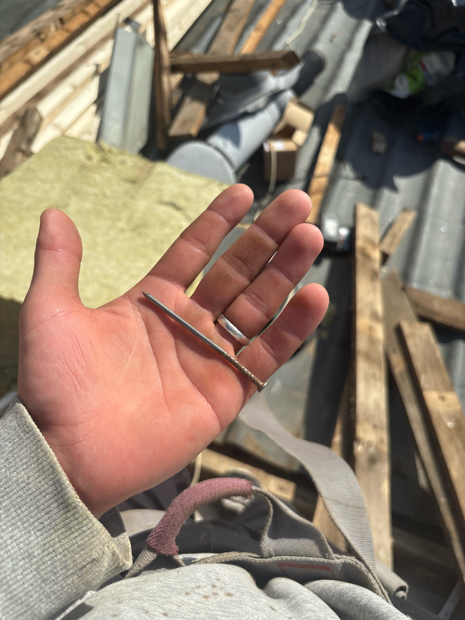 The image shows a person's hand holding a nail. The hand is pink and appears to be clean. The nail is silver and has a pointed end. The background is blurred, but it appears to be a construction site with wooden planks and other building materials. There is no specific information about the location in the city of Zhavoronki, Russia, or any details about people, subjects, objects, actions, or interactions.