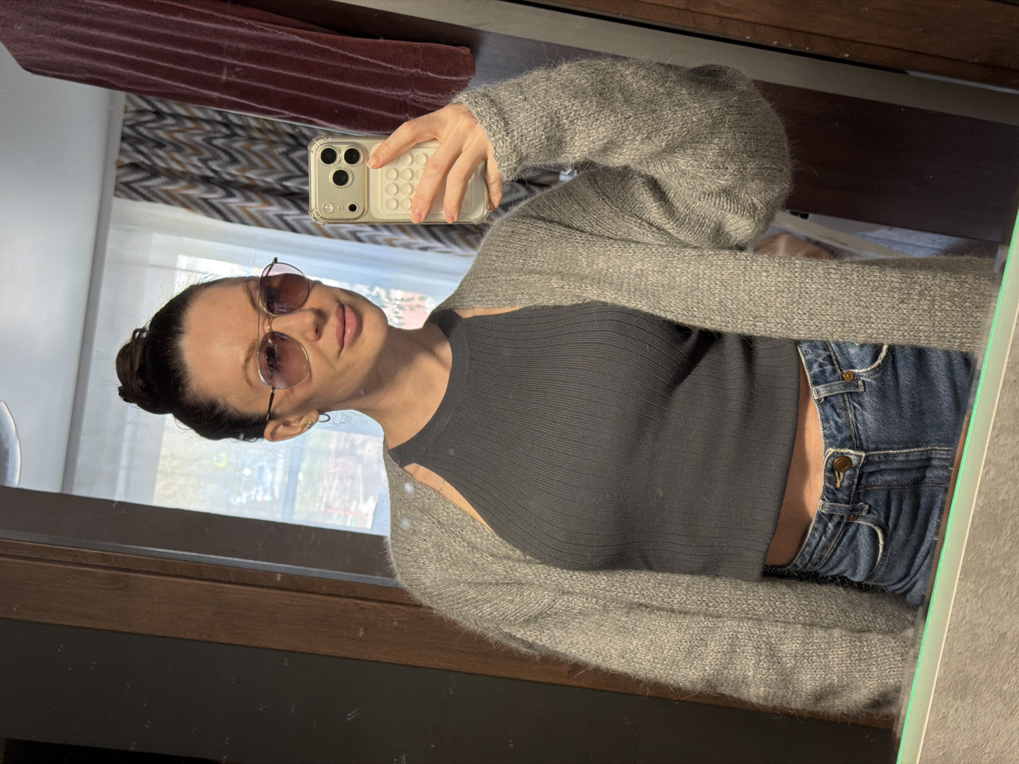 A woman wearing sunglasses, a gray knit cardigan, and a gray top is taking a selfie in a mirror. She is wearing blue jeans. The mirror reflects a room with a window and various objects, including a curtain and wooden furniture. It's not possible to identify the location in the city of Perm, Russia, based on the image.