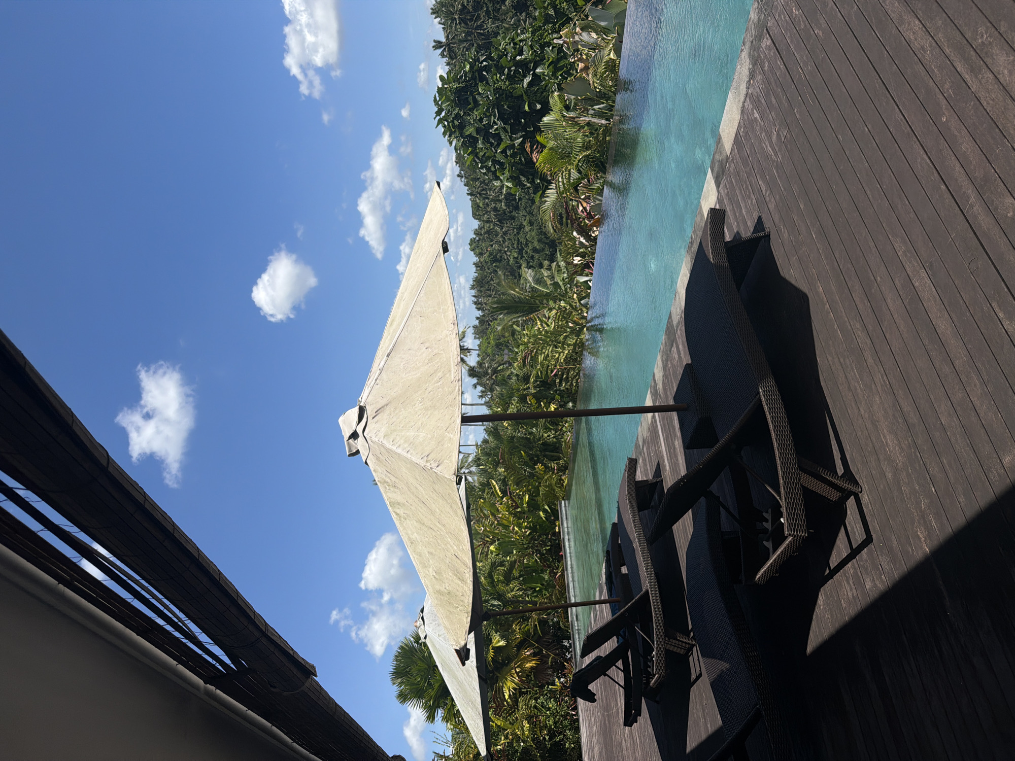 The image depicts an outdoor setting in Petulu, Indonesia, likely a resort or private residence. A light-colored umbrella is positioned near a pool, casting shadows on the dark wooden deck. Two dark chairs are placed on the deck, also shadowed. Lush tropical vegetation is visible beyond the pool's edge. The sky is blue with scattered white clouds. A building roof is visible on the left.