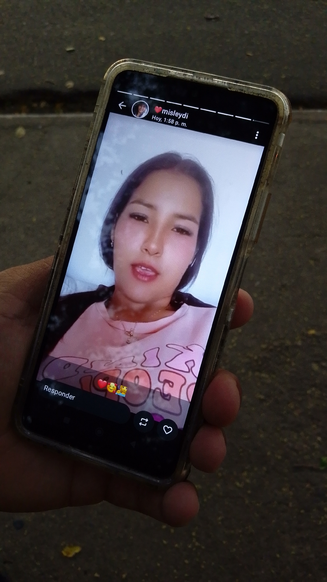 A hand holds a smartphone displaying a video of a woman. The woman in the video is wearing a pink sweatshirt with cartoon text, a gold necklace, and has shoulder-length black hair. The smartphone screen also displays Instagram interface elements such as a username "@misleydi" with a heart symbol, a timestamp "Hoy, 1:58 p.m.", and interactive icons for replying, sharing, and liking. The background suggests an outdoor location, likely a paved area in an urban setting. The city of Tinaquillo, Venezuela cannot be determined from this image.