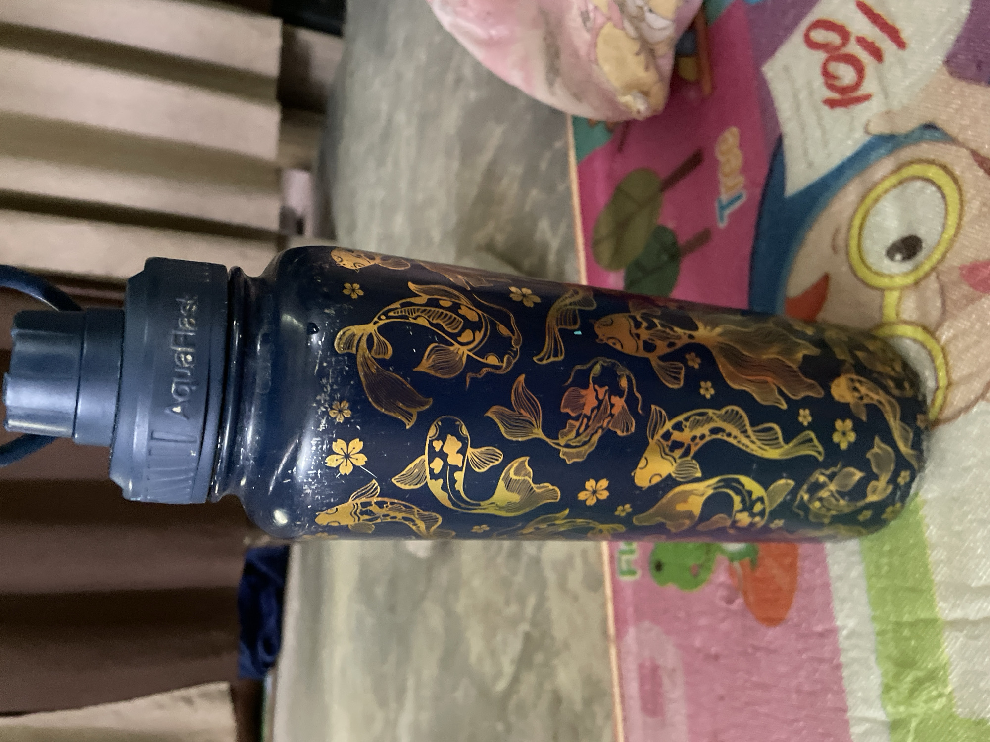 A close-up, slightly high-angle shot shows a dark blue water bottle adorned with golden koi fish and flower designs. The bottle is lying horizontally on a surface that appears to be a children's playmat with cartoon figures and bright colors. The brand name "AquaFlask" is visible on the blue cap of the bottle. The background includes a glimpse of light-colored wooden slats and a partially visible object covered in pink fabric. The image was taken indoors. Hijo, Philippines cannot be determined from this single image.
