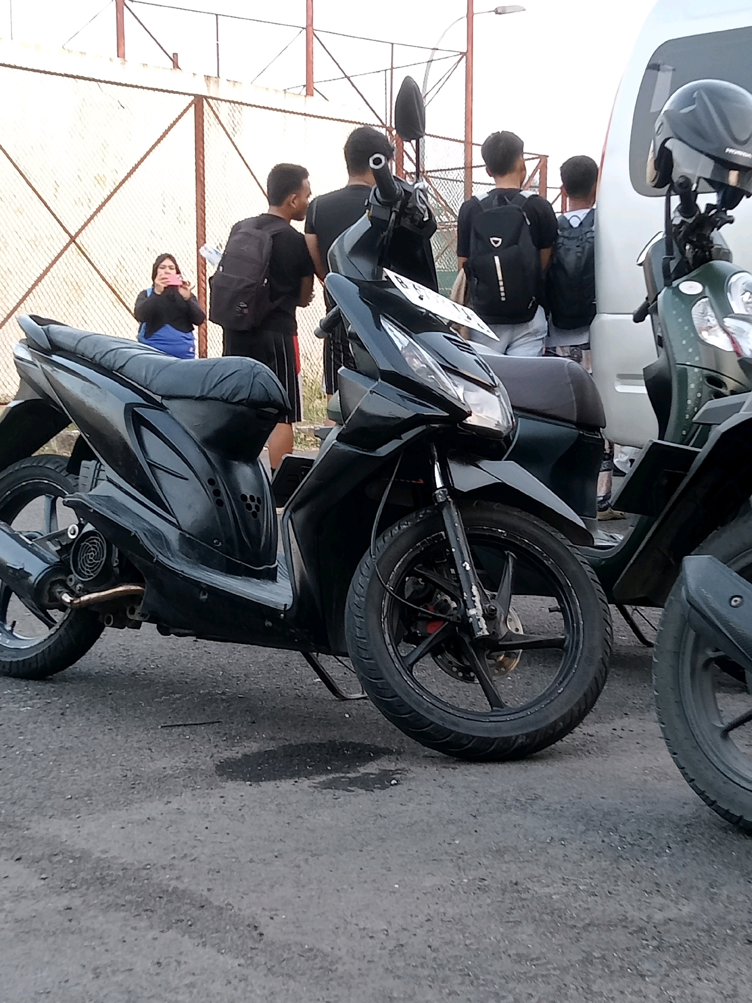 In Karawang, Indonesia, a group of individuals stands near parked motorcycles in front of a chain-link fence and a light-colored van. A woman in a blue top is holding a pink phone, appearing to take a photo or video. Two young men, one wearing a black t-shirt and shorts with a backpack, and another with a dark backpack, stand facing away from the camera. A third person, possibly male, in a dark t-shirt and shorts, is visible behind them. The foreground is dominated by two scooters, one black and one dark green, with their front wheels and parts of their frames visible. The ground is paved asphalt.