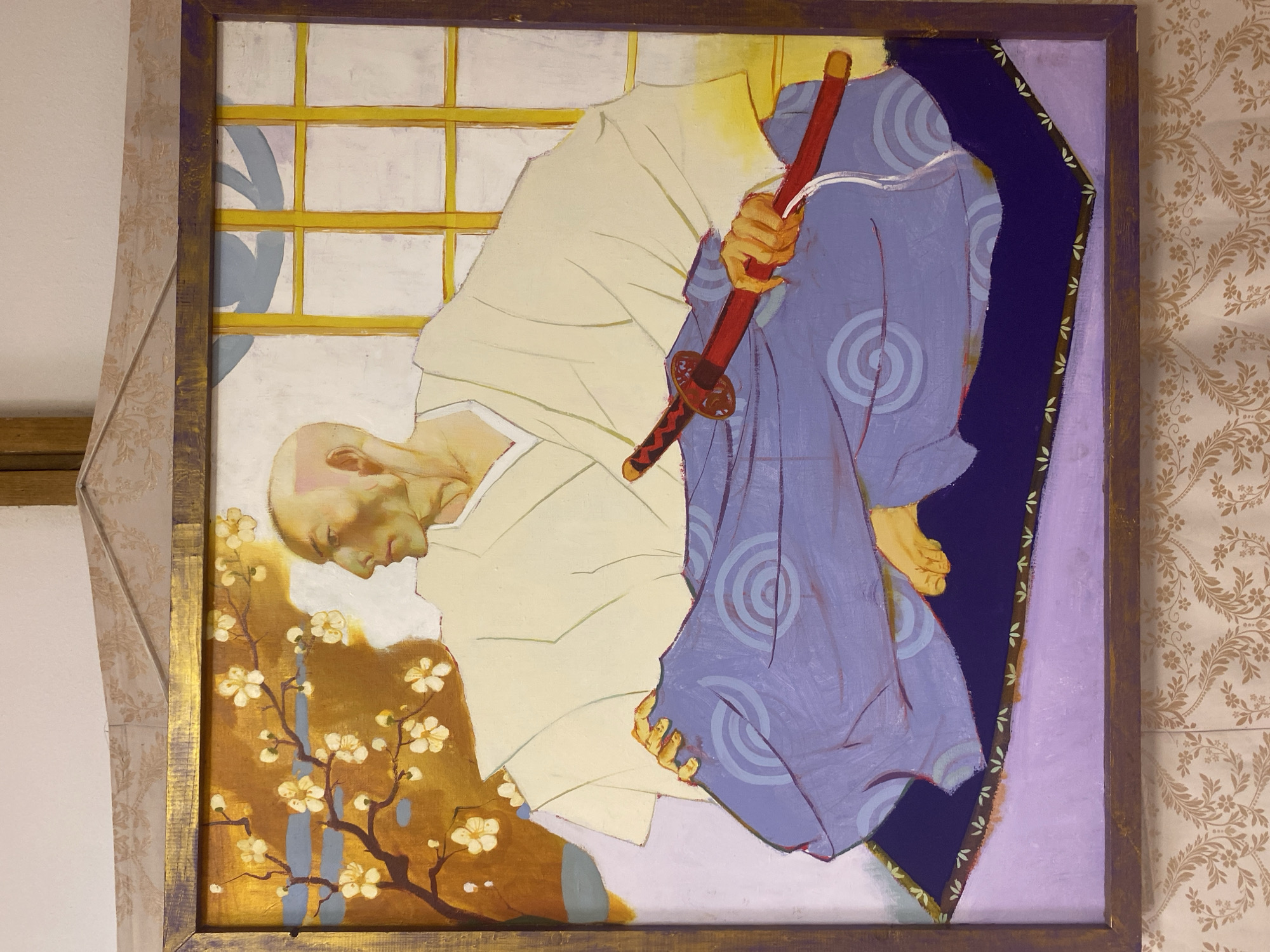 The image shows a painting of a bald man with a pale complexion and sharp features, depicted in profile, looking to the left. He is wearing a cream-colored robe with wide sleeves and a purple garment patterned with concentric circles. The man is seated on a dark purple cushion with a lighter purple base, resting his feet on the cushion. His right hand holds the hilt of a red katana, while his left hand rests on his lap.
Behind the man, a window with yellow panes is visible. To the left of the man, a branch with white flowers and dark brown stems is depicted against an orange and blue background. The painting is framed by a rustic wooden frame with a metallic sheen. The painting is hung on a wall adorned with a patterned wallpaper. The overall scene suggests an indoor setting, possibly a traditional Japanese room. There is no information about the location of this image within a city.