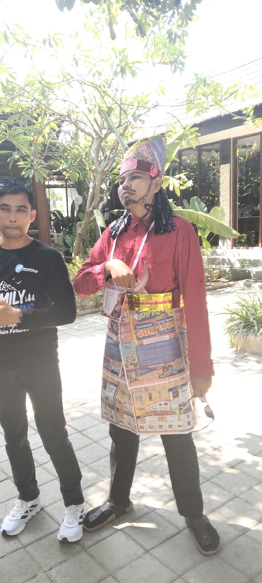 In Talise, Indonesia, two individuals stand outdoors under dappled sunlight filtering through leafy trees. The person on the right is dressed in a striking costume, featuring a red long-sleeved shirt, black pants, and an elaborate headdress adorned with colorful patterns. A faux beard and mustache are visible, and a sheathed dagger is tucked into a yellow and gold sash around their waist. This sash is made of what appears to be folded newspaper or magazine pages, depicting various advertisements and text, including mentions of "CASHBACK 50K" and "UANG TUNAI." A name tag or lanyard with the name "SULTAN HASANUDDIN" and a portrait is also attached to the costume. The person on the left wears a black long-sleeved t-shirt with a graphic design featuring a mountain and the word "FAMILY," along with black pants and white athletic shoes. Both individuals are standing on a paved walkway made of square tiles. In the background, there's a building with large glass windows and lush green foliage, suggesting an outdoor setting possibly near a recreational or event space.
