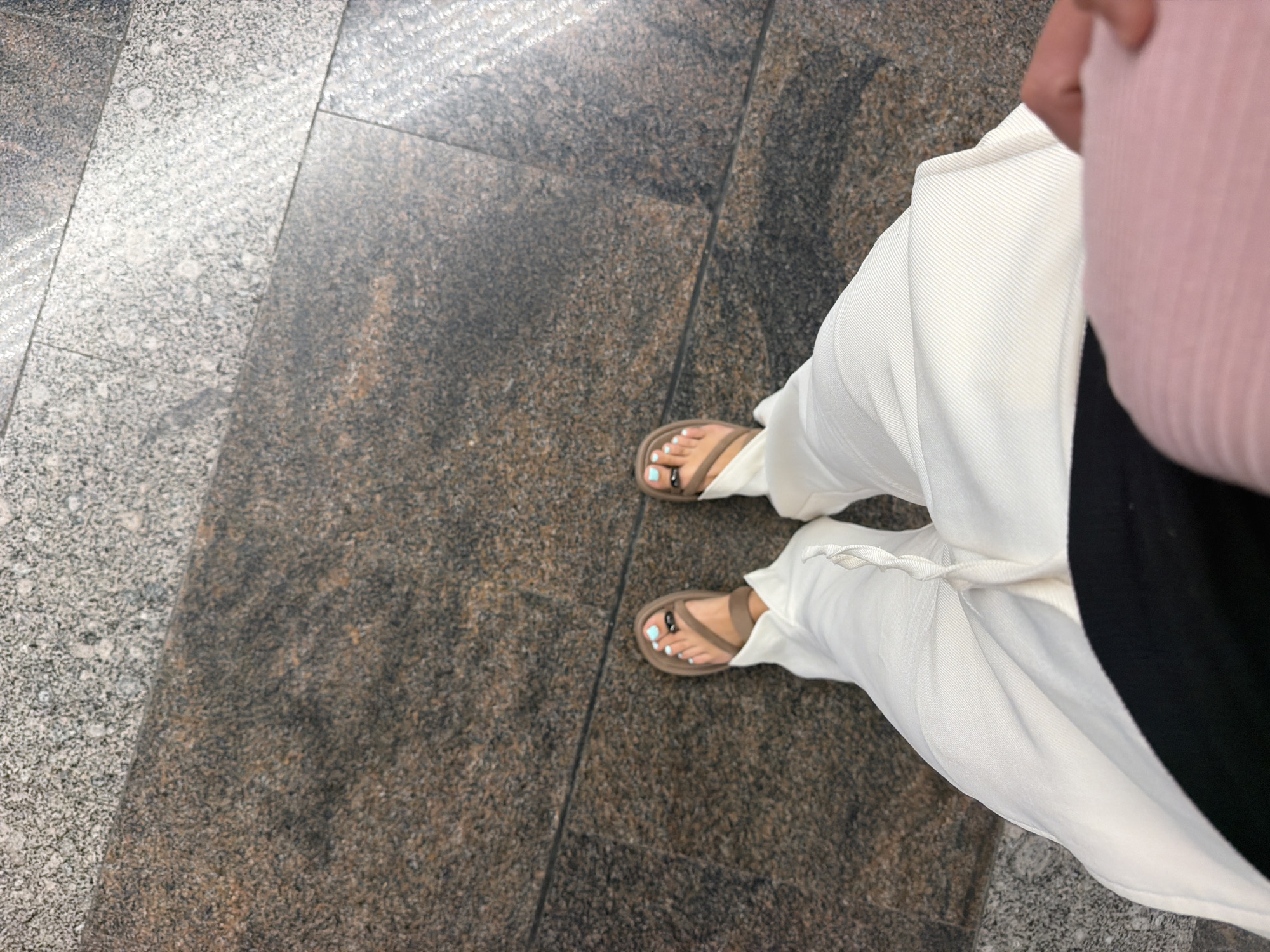 A first-person perspective shot captures a pair of feet in light-colored sandals and white pants, standing on a granite-tiled floor. The sandals are brown with a simple strap design, and the toenails are painted a light blue. A black band is visible on one toe. The floor is composed of large, dark, speckled granite tiles with lighter granite tiles also present. The lighting suggests an indoor or shaded outdoor location. No other individuals or significant environmental features are visible. The context of Sochi, Russia, is not visually apparent within the image itself.