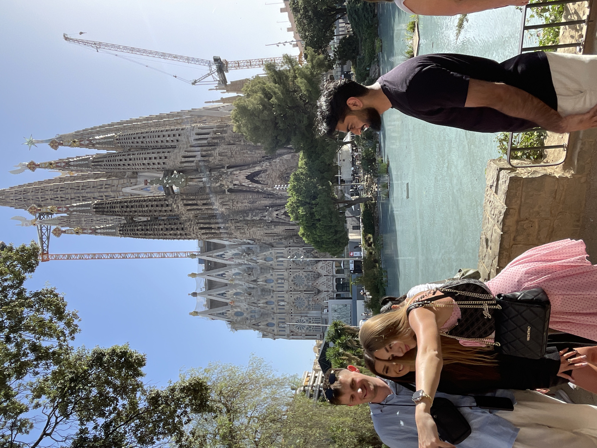 In Barcelona, Spain, at the Sagrada Familia, a group of four people are captured in a photograph. The iconic basilica, still under construction with cranes visible, dominates the background. In the foreground, a man in a light blue shirt is taking a selfie with two women. One woman, wearing a pink polka-dot dress, is holding the phone and smiling, while another woman in a black dress stands beside her. To the right, a man with a beard in a black t-shirt faces away from the camera, looking out at the scene. A body of turquoise water is visible in the midground, reflecting the blue sky. Lush green trees and foliage surround the area, adding to the vibrant atmosphere.