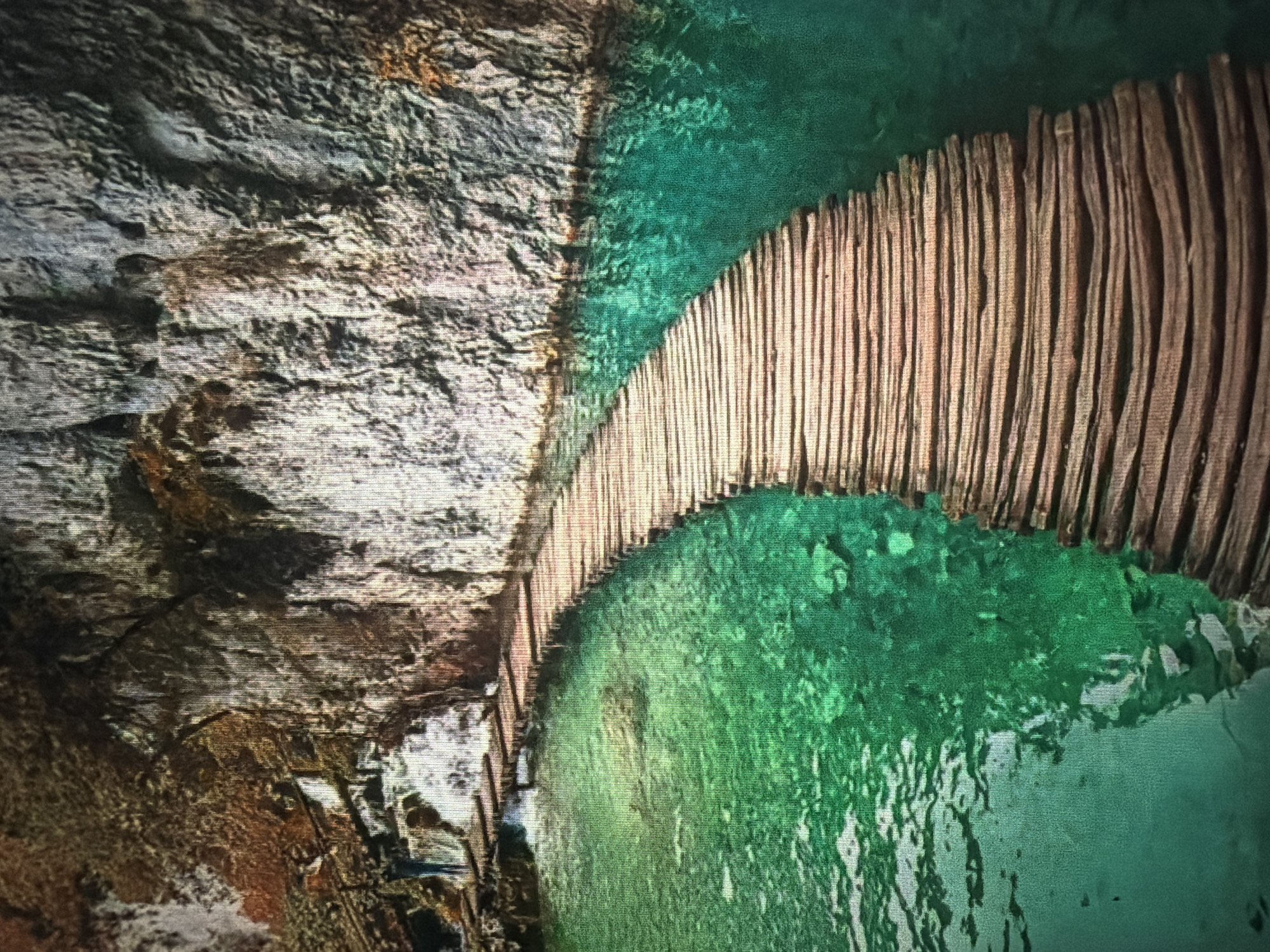 The image shows a wooden boardwalk winding through a turquoise lake, bordered by a sheer rock face and lush vegetation. A single person stands on a section of the boardwalk near a small waterfall. The water is clear, revealing submerged rocks and the lakebed. The boardwalk is constructed of rough-hewn wooden planks, forming a curving path that extends into the distance. The rock face is textured and variegated in color, with patches of moss and vegetation growing on its surface. The overall scene evokes a sense of natural beauty and tranquility. There is no information in the image that indicates this location is in the city of Baan Mae Nam, Thailand.