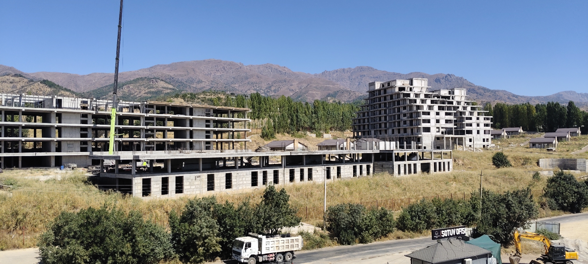 This is a wide-angle, panoramic photograph taken on a clear day in Aktash, Uzbekistan, depicting a construction site with several large, unfinished buildings against a backdrop of arid, mountainous terrain. The primary focus is on two multi-story concrete structures in various stages of construction, characterized by exposed frameworks, empty window openings, and concrete floors. A large construction crane, partially obscured by the buildings, is visible on the left side of the frame. In the foreground, a white dump truck is parked on a paved road, and a yellow excavator is situated near a small, dark-colored building with a green canopy. Several smaller, completed residential-style buildings with dark roofs are scattered on the hillside behind the main construction. The environment is dry and sparsely vegetated with grass and scattered trees. The sky is a clear, bright blue.