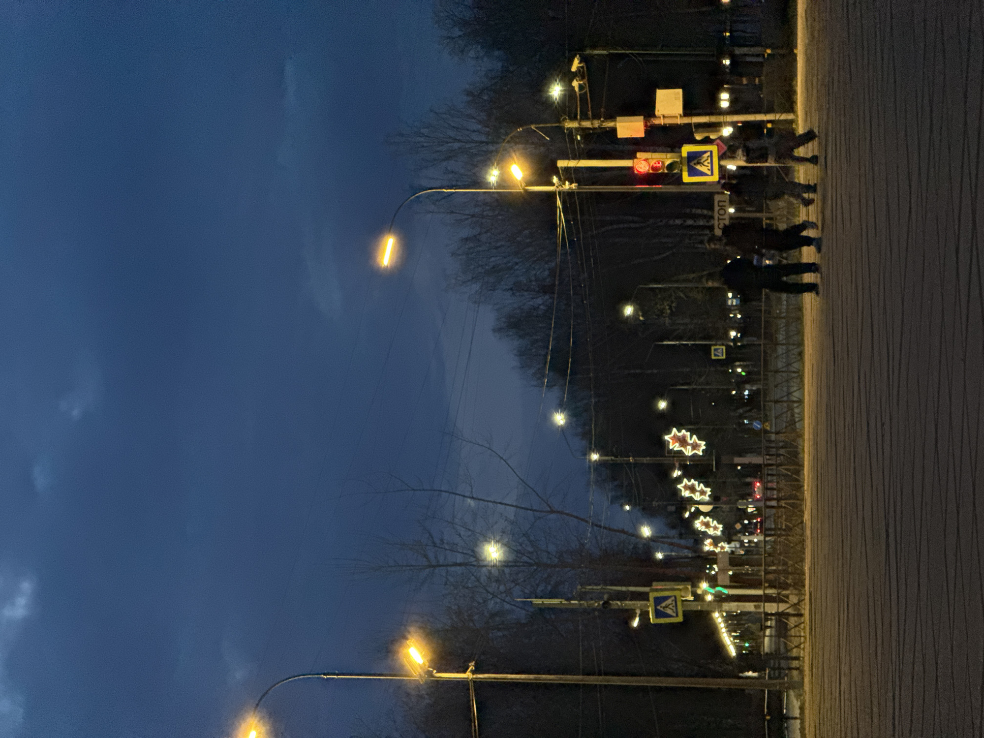 The image captures a nighttime street scene in what appears to be Chaikovsky, Russia, given the "СТОП" sign and the street layout. The foreground features a wide sidewalk or pedestrian area with vertical lines, likely indicating paving slabs. Streetlights with yellow illumination line the street, and the sky is a deep blue. There are bare trees between the sidewalk and the street, with decorative star-shaped lights strung between some of the trees.
In the distance, a pedestrian crossing with a stop sign is visible. A traffic light is red. Several individuals are walking along the sidewalk toward the crosswalk. There are utility poles with wires running along the street.