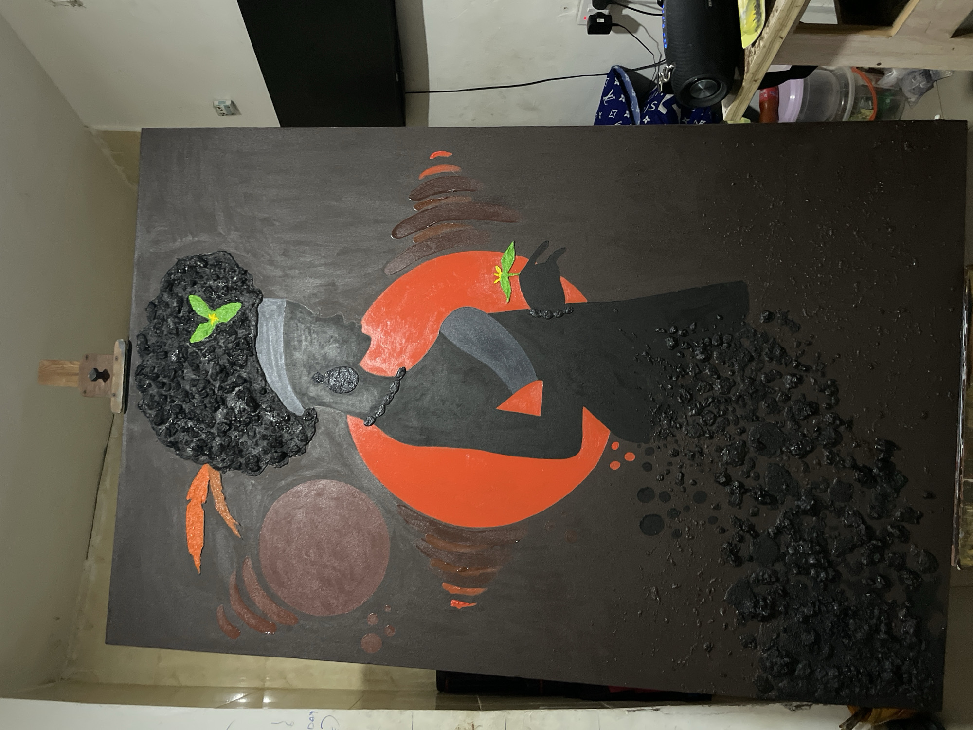 A close-up, eye-level shot shows a large, rectangular painting leaning on an easel within an indoor space. The painting features a dark background with abstract shapes and figures. A red circle is prominent, with dark, textured materials used to depict a head and body in profile. There are also other shapes and textured elements, including a dark sphere and stacked horizontal lines. The background is a light-colored interior wall. The location is in Ado, Nigeria.