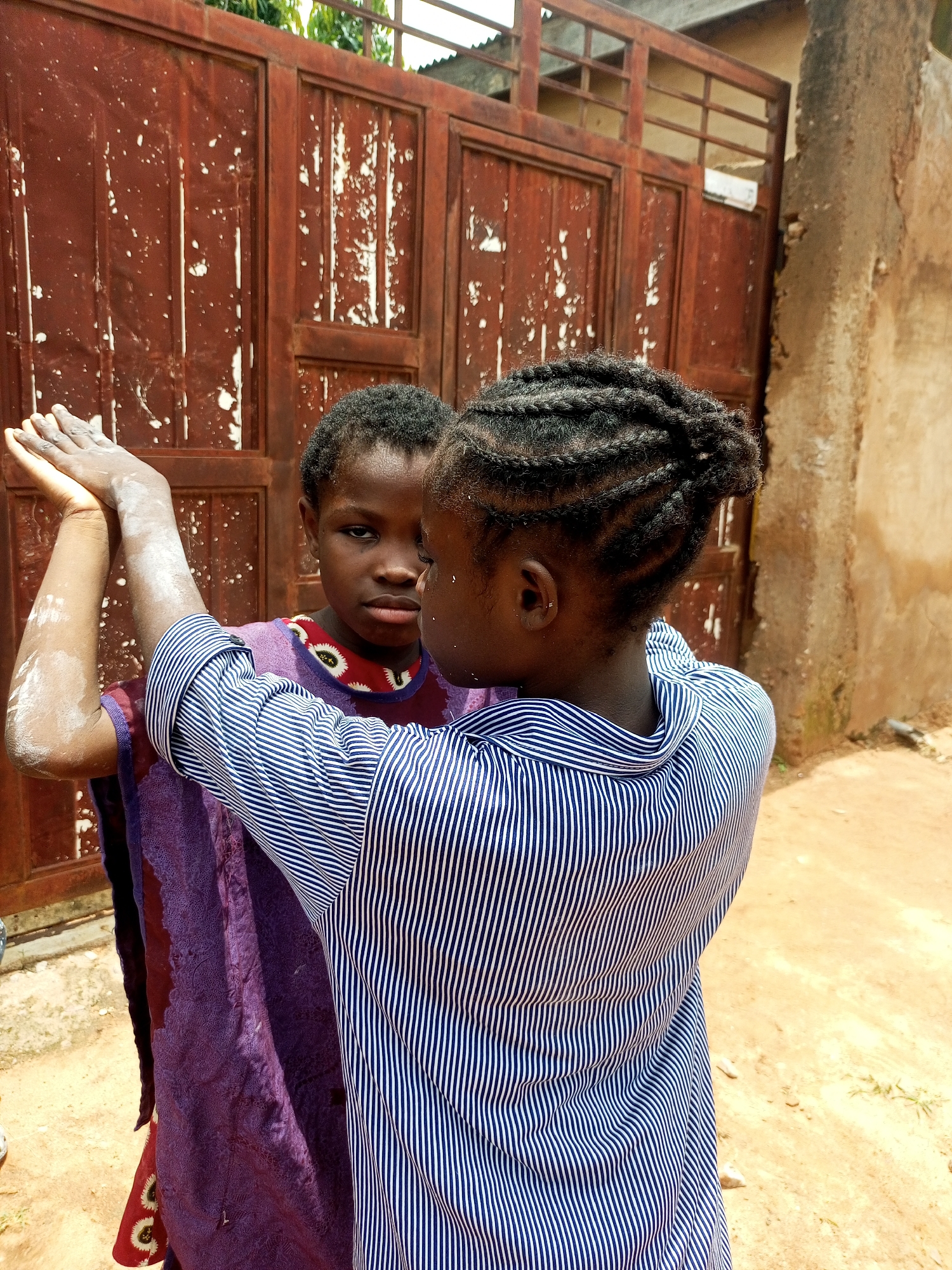 Two young girls stand in front of a weathered, reddish-brown metal gate with peeling paint, likely in Tempure, Nigeria. The girl on the left, facing forward, has dark, short hair and is wearing a purple garment with a red and white patterned trim. Her arms are raised, and her hands are positioned as if in a clapping motion, with white powder dusted on her arms and hands. The girl on the right, seen from behind, has her hair styled in intricate braids. She is wearing a blue and white striped shirt and is facing the girl on the left, her arms also raised and extended towards the other girl. The ground beneath them is sandy and appears to be unpaved. Behind the gate, a building with a tan, textured wall is visible. The overall impression is of a candid moment captured outdoors.