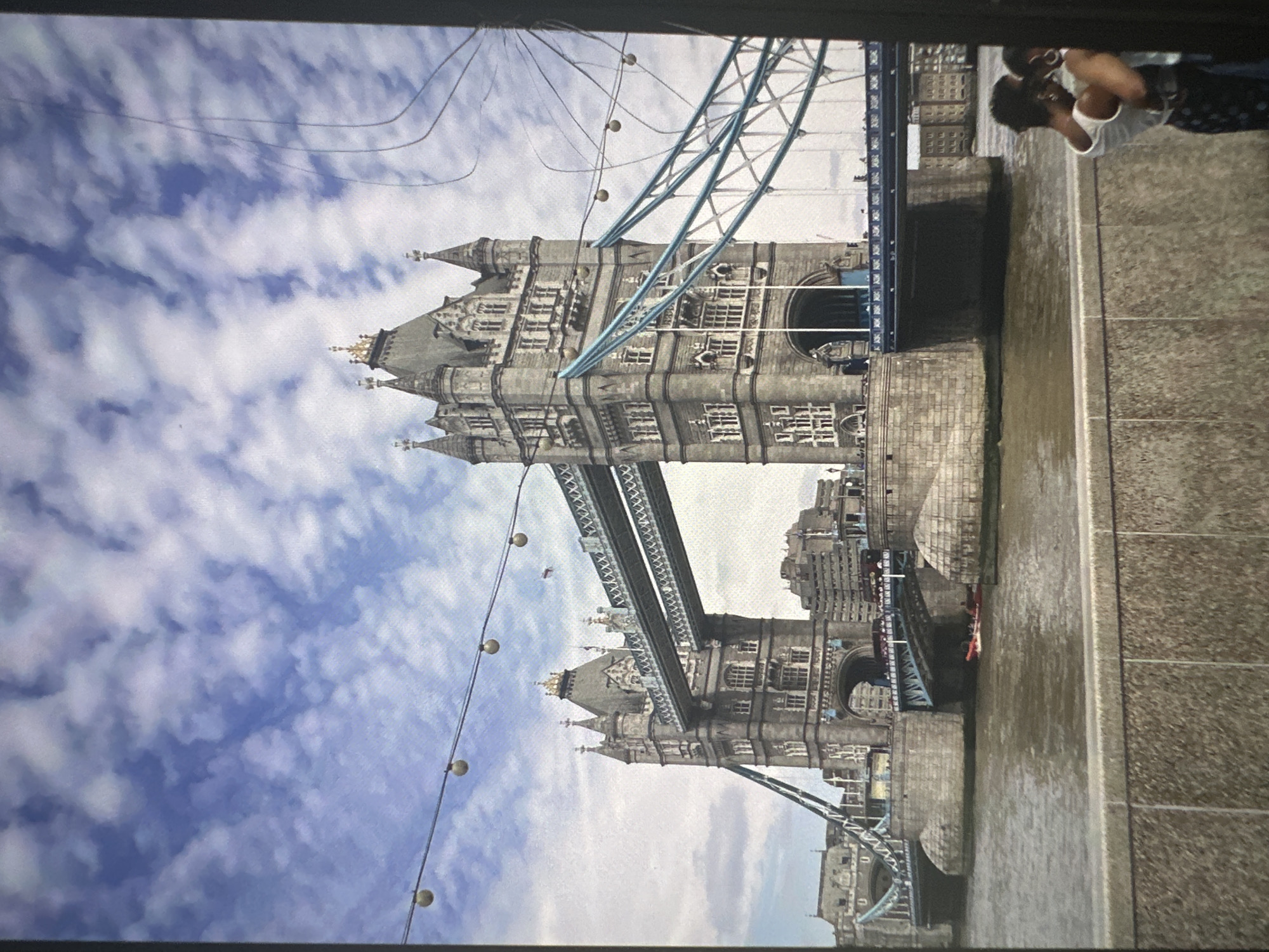 The image shows Tower Bridge in London, United Kingdom, under a cloudy sky. The bridge is a bascule and suspension bridge that crosses the River Thames. It is made of stone and steel and features two towers. The bridge is a popular tourist attraction and is often seen with boats passing underneath. In the foreground of the image, two people are standing on a stone wall, facing away from the camera. They appear to be a couple. The river is visible below the bridge, with a small red boat on it. The image is taken from a slightly elevated position, looking towards the bridge. The overall impression is of a busy, iconic landmark on a pleasant day.