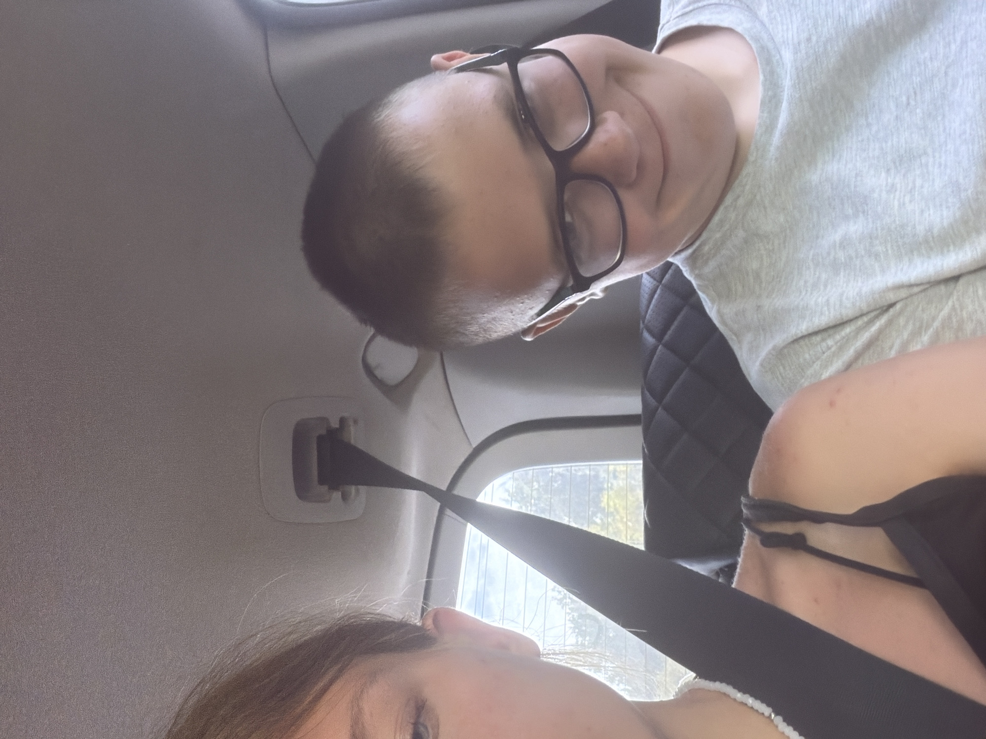 Two people are in the back seat of a vehicle. On the left, a young female's face is partially visible, looking towards the camera. She has blonde hair pulled back and is wearing a black tank top and a beaded necklace. A black seatbelt is diagonally across her body. On the right, a young male wearing glasses and a grey t-shirt smiles at the camera. The interior of the vehicle is visible, with leather seats and a seatbelt mechanism on the ceiling. The background outside the car window shows trees and a blue sky with clouds. The image appears to be a selfie taken inside a car.