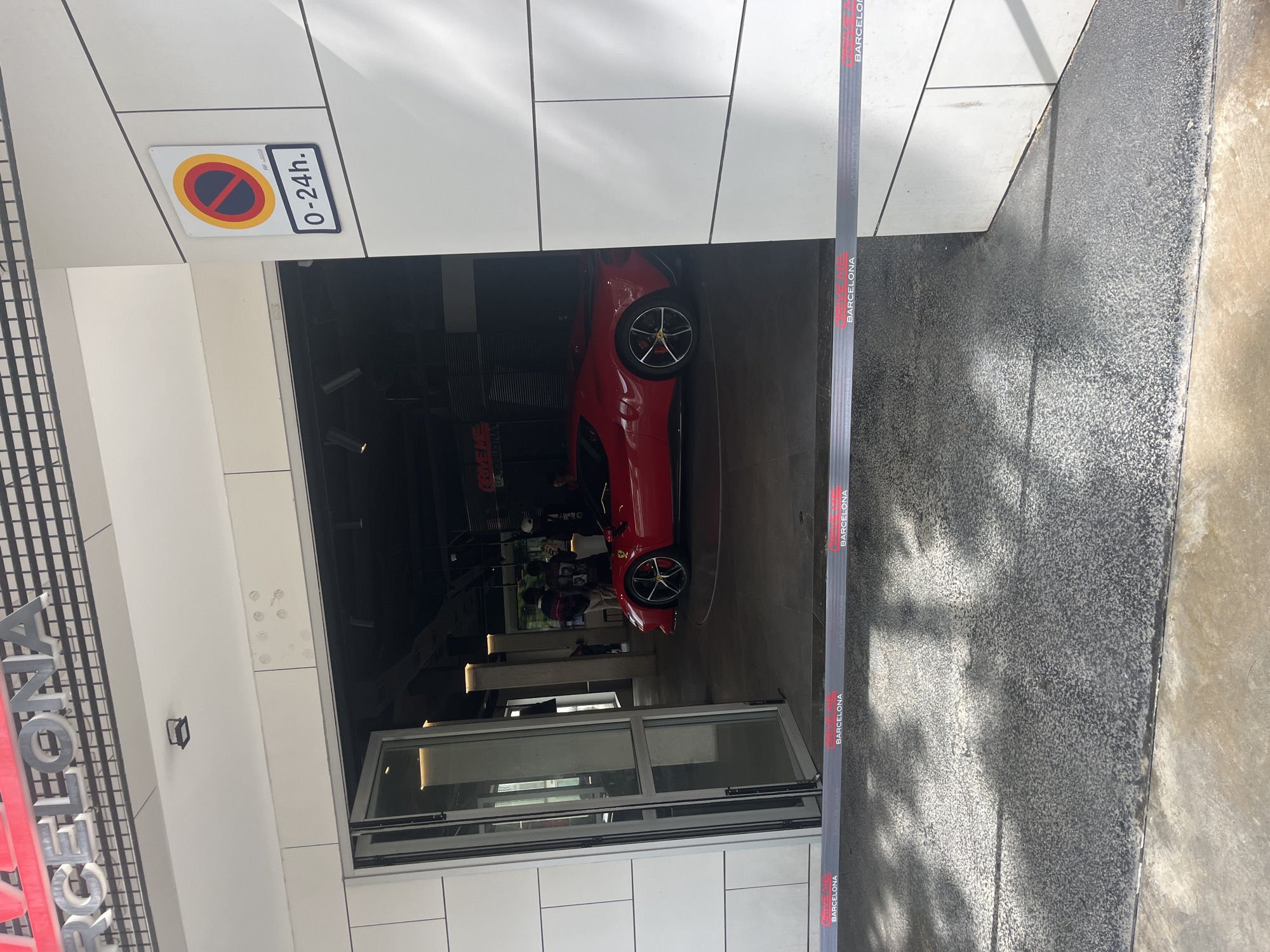 The image, oriented on its side, displays the entrance of a modern building, likely a car showroom or experience center in Barcelona, Spain. A vibrant red sports car, possibly a Ferrari, is prominently parked inside the well-lit interior. The car features black multi-spoke wheels with red brake calipers. Several indistinct figures are visible further inside, suggesting the presence of visitors or staff. The building's facade is constructed from white rectangular panels, with glass doors providing access to the interior; one door is partially open. To the upper left (when viewed upright), a white sign with a "No Stopping/No Parking" symbol indicates a "0-24h." restriction, signifying 24-hour no parking. A black barrier tape spans vertically across the right side of the frame, repeatedly emblazoned with "DRIVE EXPERIENCE BARCELONA" and "BARCELONA" in red and white text. On the far left, large metallic letters, partially obscured, spell out "DRIVE EXPERIENCE BARCELONA", further establishing the venue. The ground leading up to the entrance is a dark, textured surface, possibly a ramp. The overall impression is that of a premium automotive experience, likely during daytime.