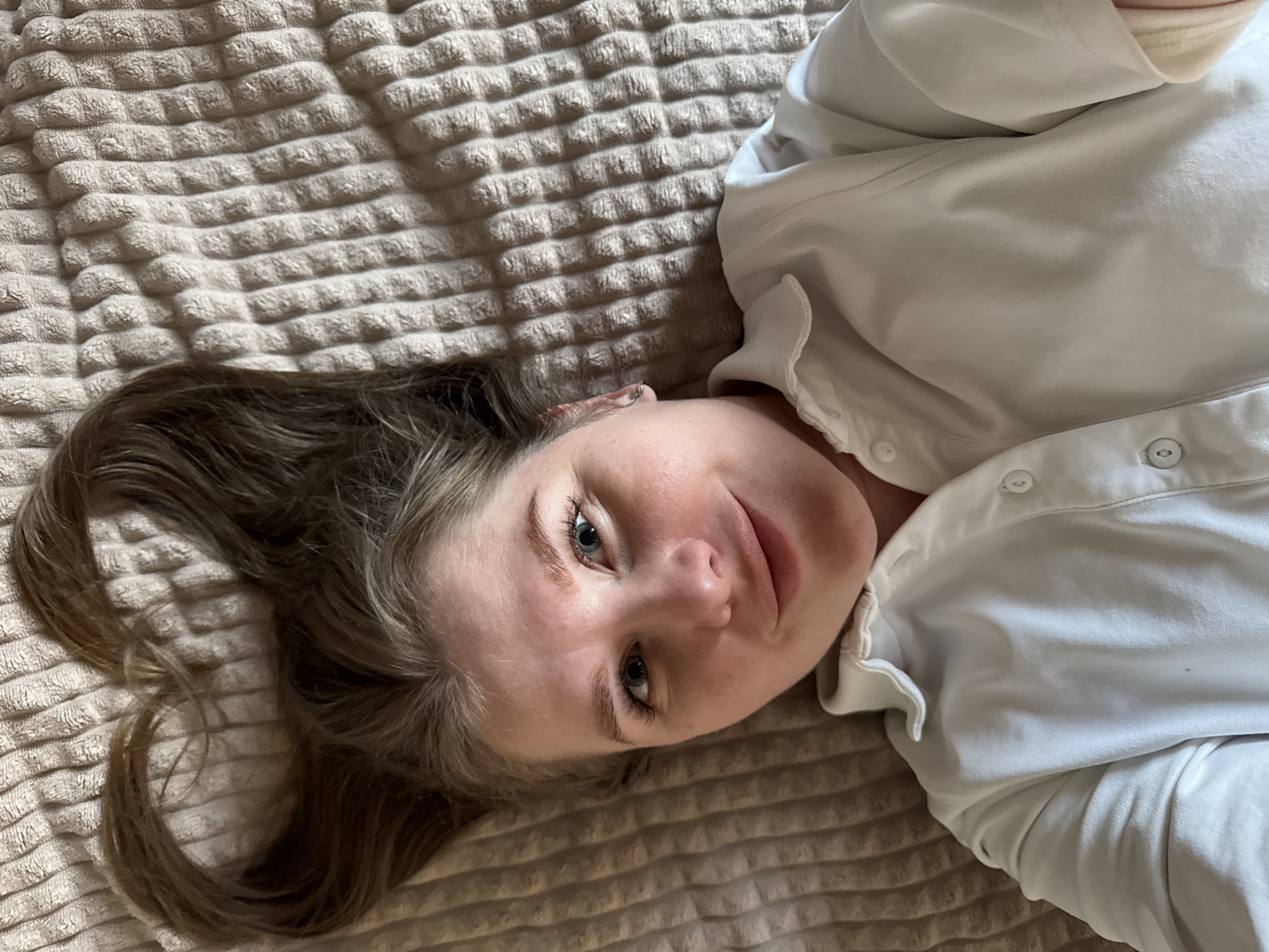 A young woman with light brown hair and blue eyes is lying on a beige textured bedspread. She is wearing a white button-down shirt. The photograph appears to be taken from an overhead perspective. The image doesn't provide enough information to discern the location in Gornyi (Dvurechenskoe S/P), Russia.
