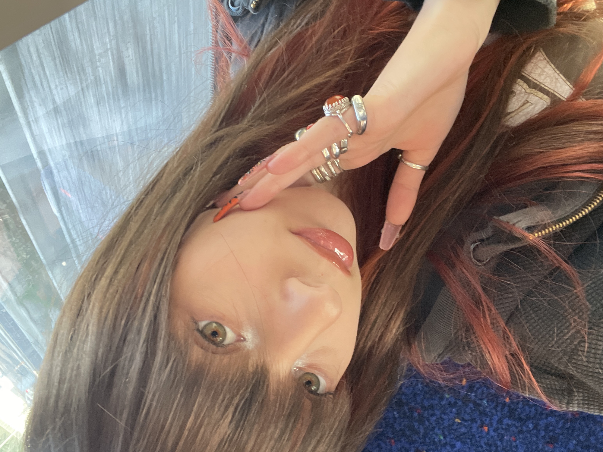 A person with auburn hair and several rings on their hand is sitting on a blue seat with orange dots, possibly on public transportation in Tel Aviv, Israel. The person's face is visible, and they have light skin and hazel eyes. The background shows a window with a blurry view of the city. The person appears to be wearing a jacket. No specific location within Tel Aviv can be determined from the image.
